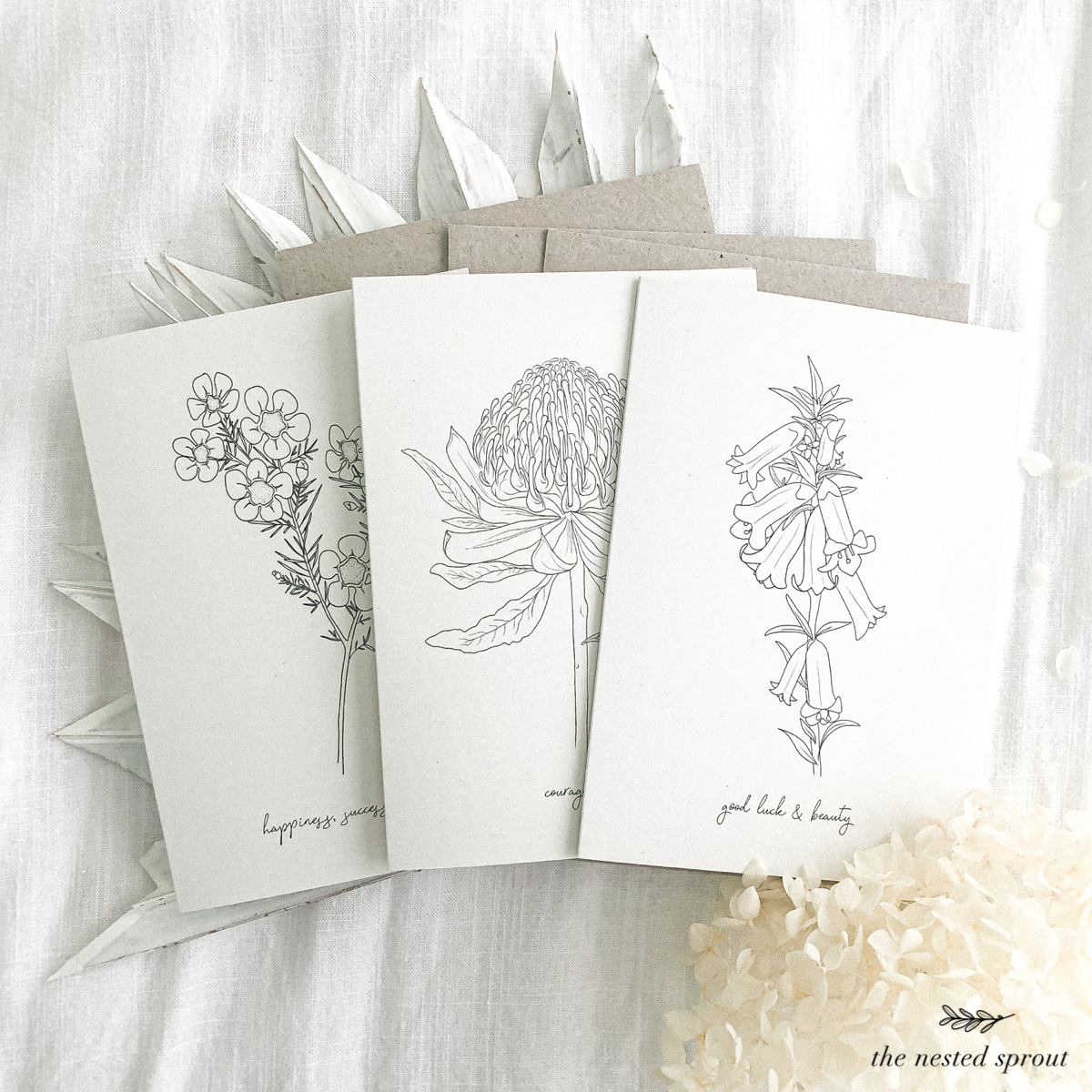 Three botanical-themed greeting cards with line art illustrations of flowers, each with a message: "happiness & success," "courage," and "good luck & beauty." The cards are arranged on a textured white surface with dried leaves and white flowers as decoration.