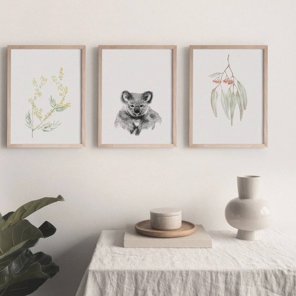 Three framed botanical illustrations hung on a white wall, with a table below holding a white vase, a bowl on a tray, and a plant in the foreground.