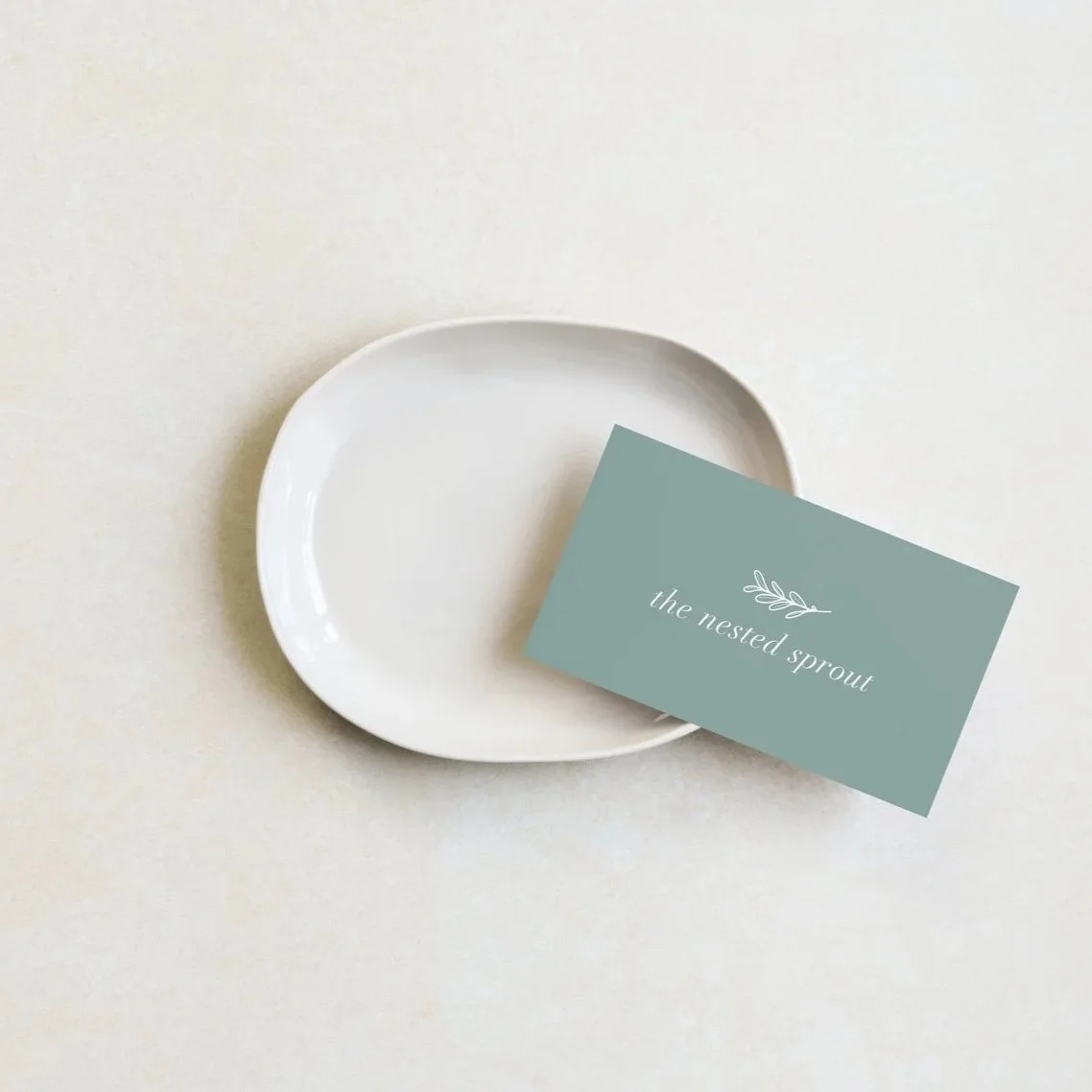 A white plate with a sage green business card that reads "the nested sprout" and has a small leaf illustration, on a light-coloured surface.