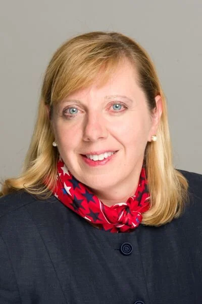 Woman with blonde hair in a navy blue jacket with a red and blue scarf around her neck.