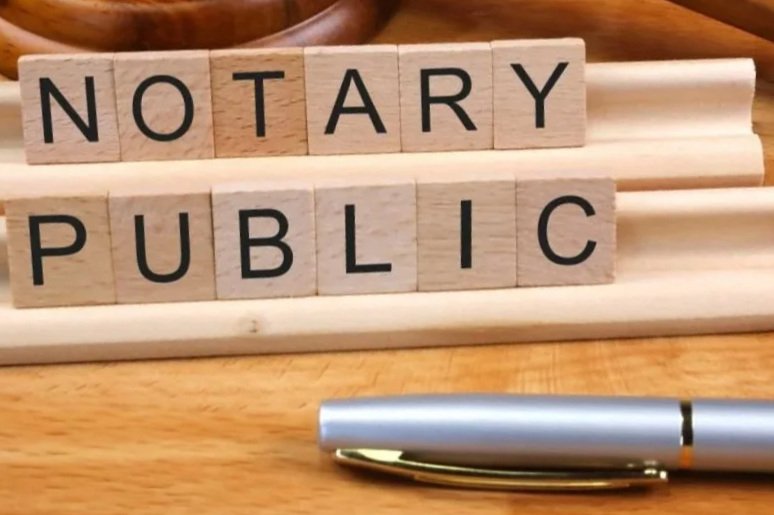 Scrabble tiles spelling 'NOTARY PUBLIC' on a wooden surface with a silver pen in front.