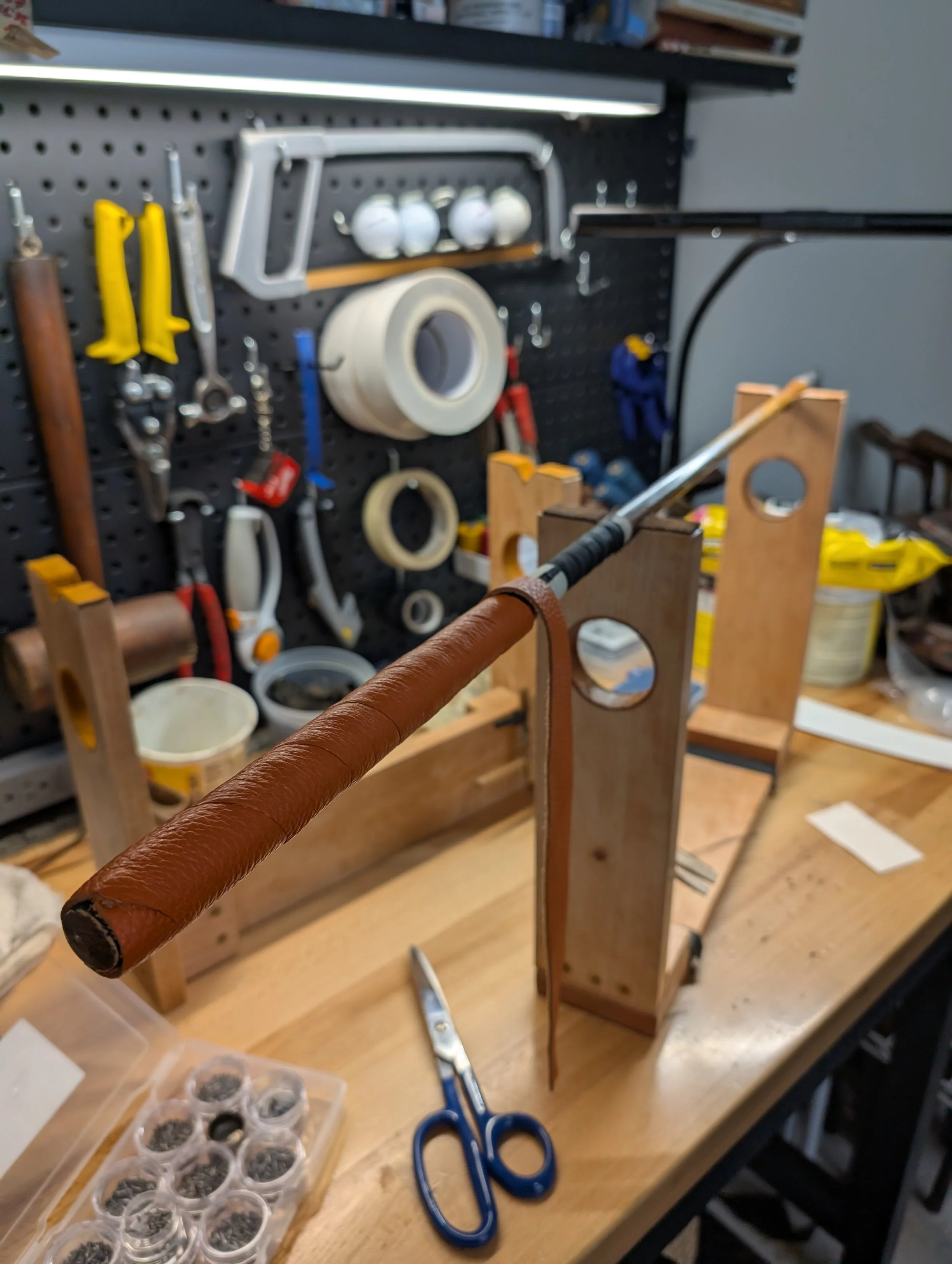 A hickory golf club refurbishment in progress on a workbench with tools and supplies in the background. A hickory golf club wrapped in a leather grip is mounted on a wooden frame, with scissors and small containers nearby.