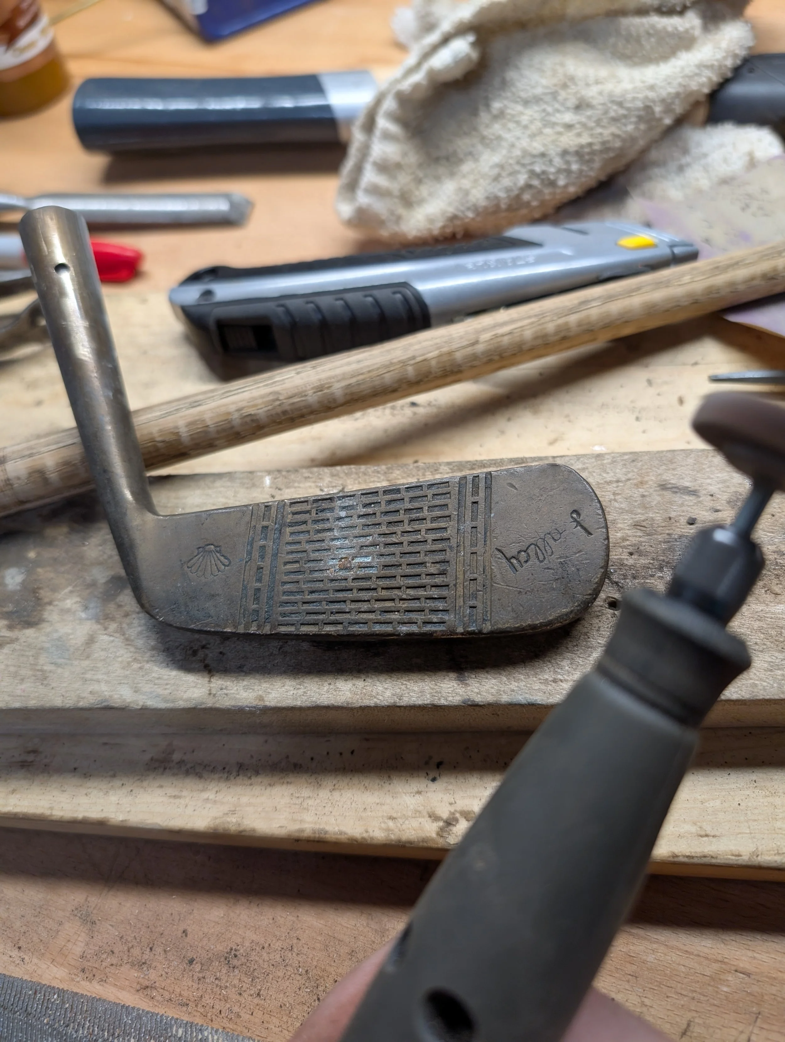 Close-up of a antique brass golf club head lying on a workbench surrounded by tools and equipment, including a utility knife, a clamp, and sanding material.