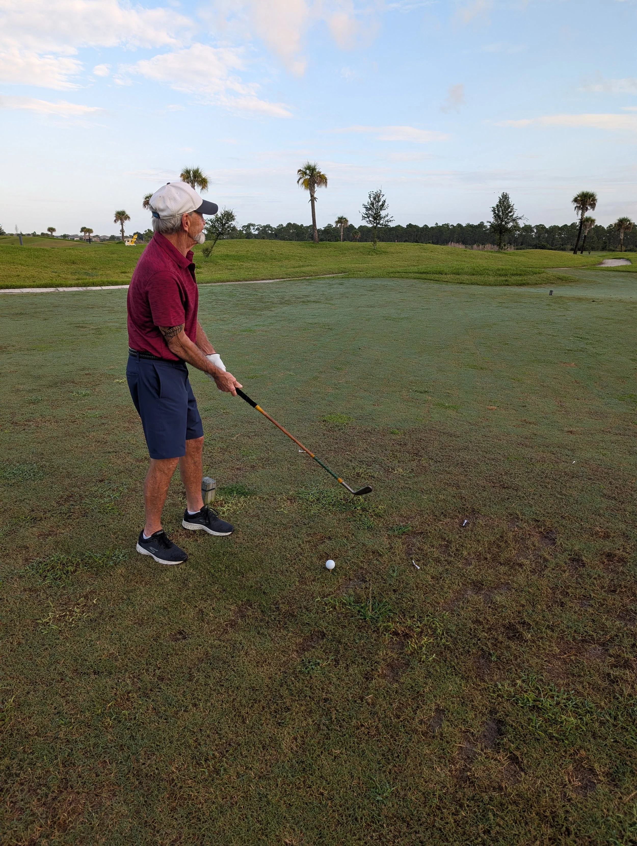 An elderly man wearing a maroon polo, navy shorts, black sneakers, and a white cap is preparing to hit a golf ball with a hickory golf club on a golf course with palm trees in the background.