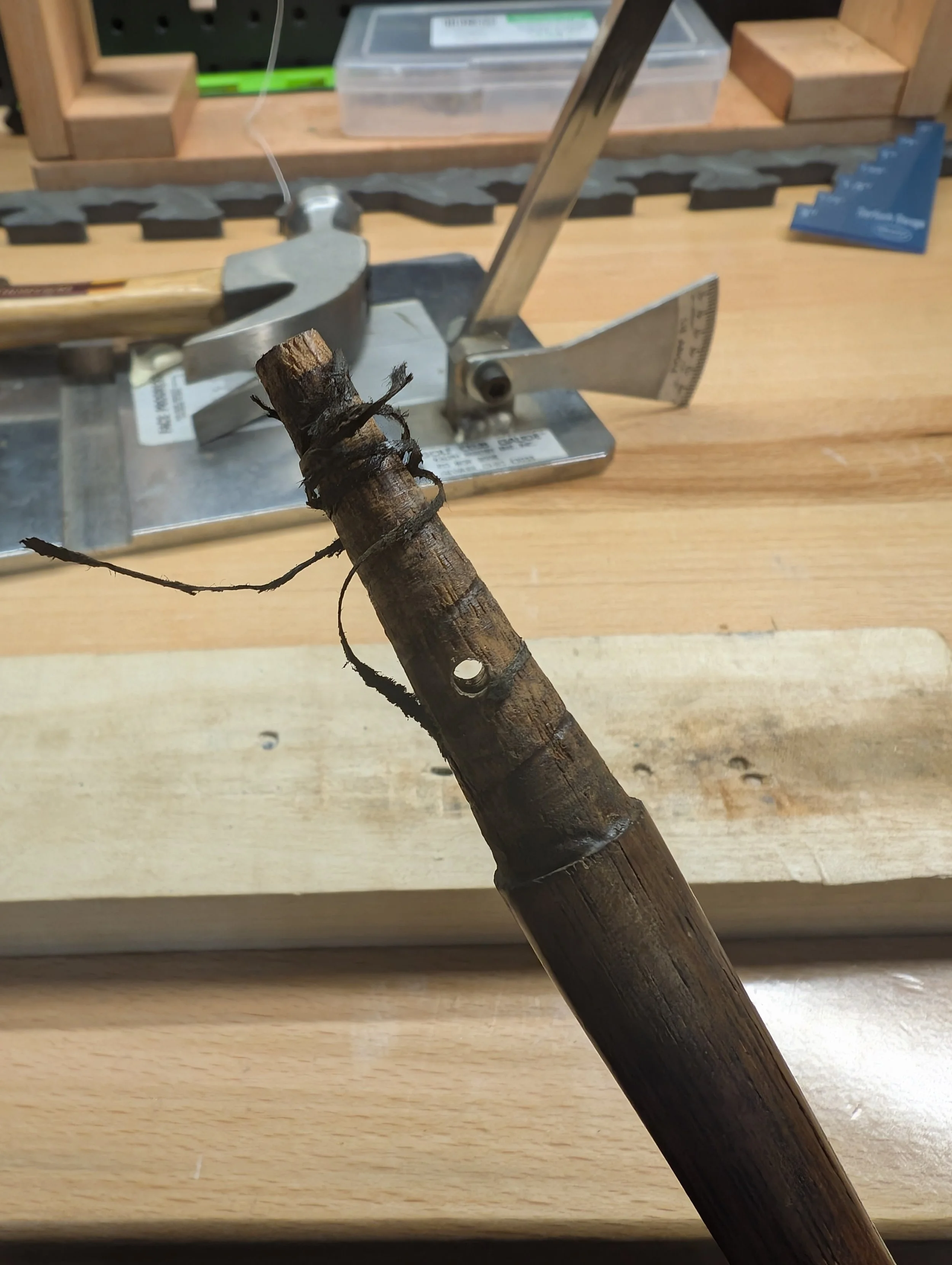 A piece of wood or a small branch with a hole drilled into it, attached with wire to a metal tool on a workbench.