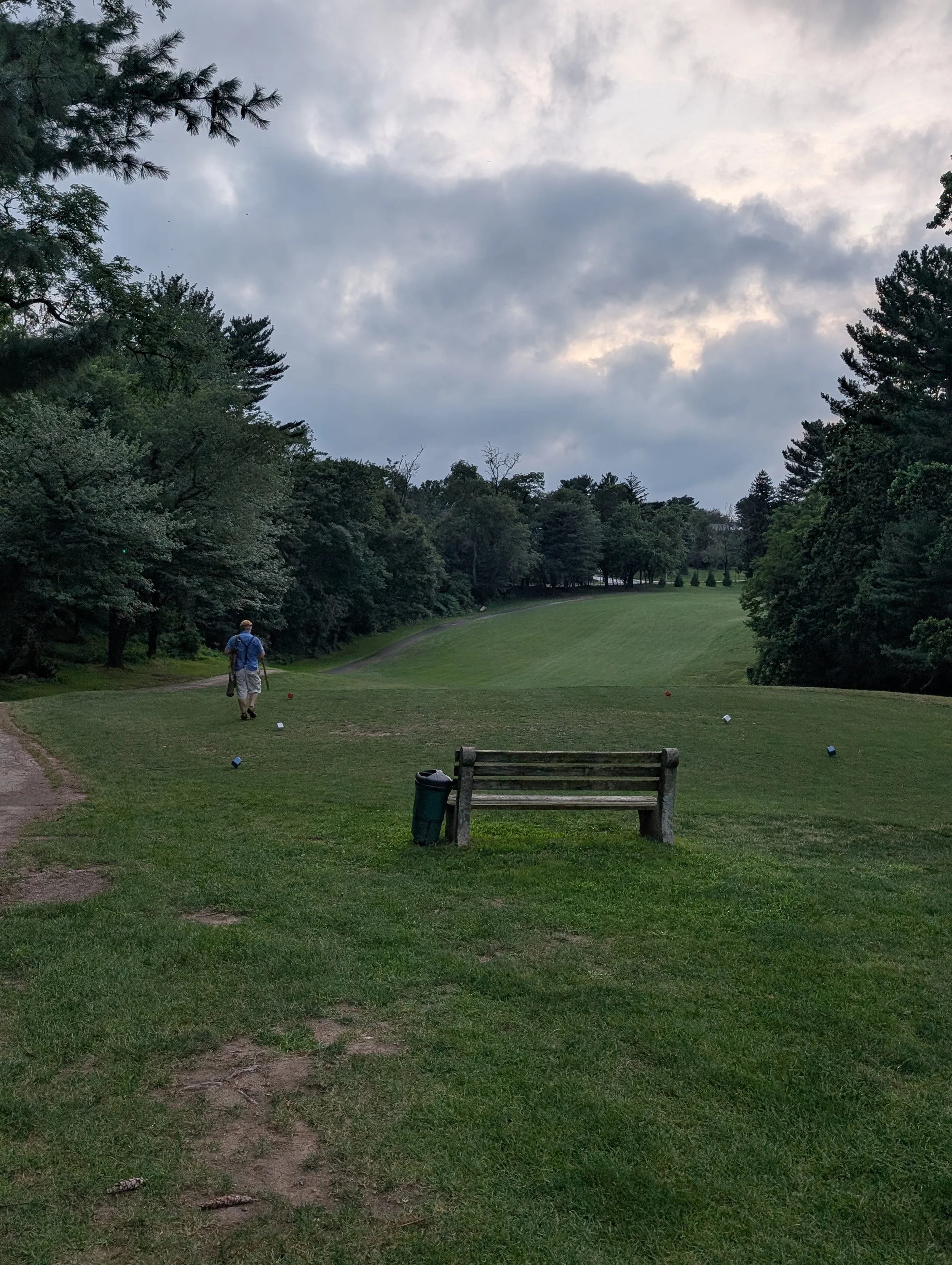 A person walking on a golf course with a hickory golf club, surrounded by trees, with a cloudy sky overhead.
