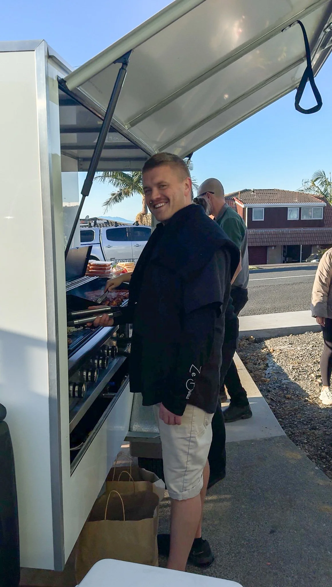 A man smiling while grilling food on a barbecue outside on a sunny day, with other people and houses in the background.