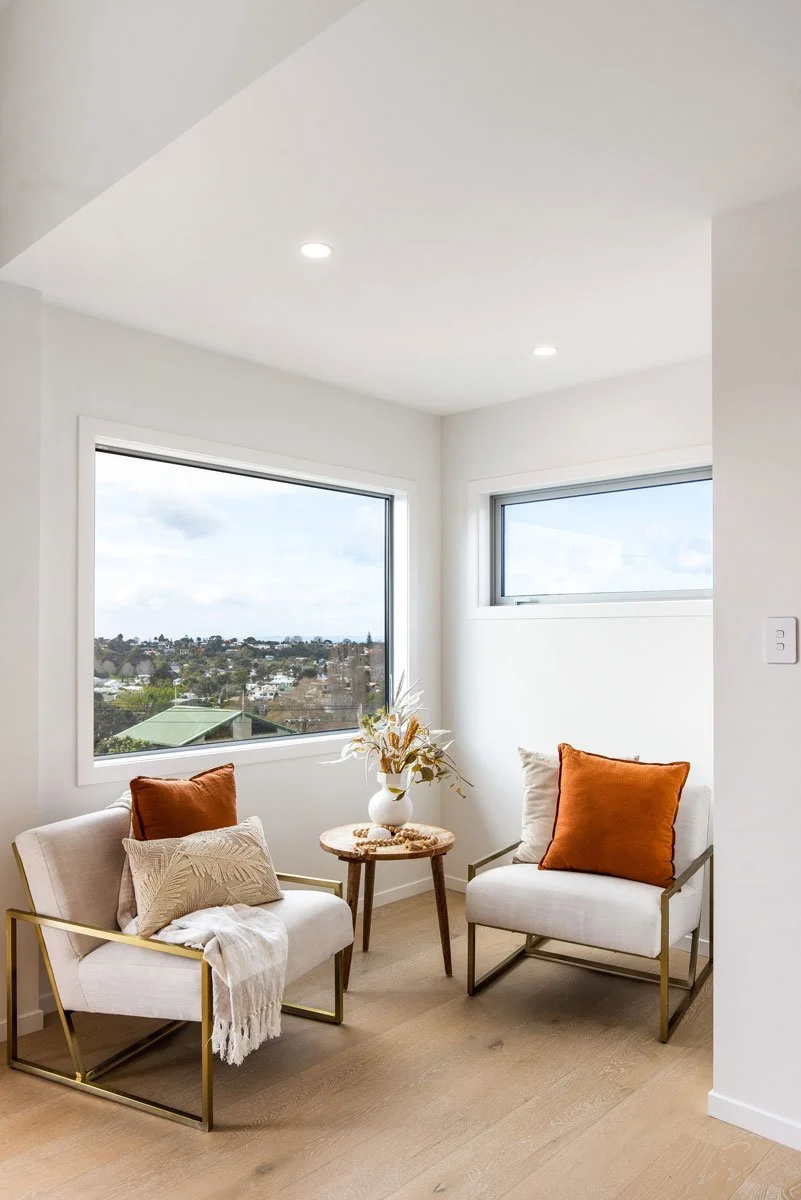 Cosy reading nook with armchairs, cushions, side table, and large windows overlooking coastal views.