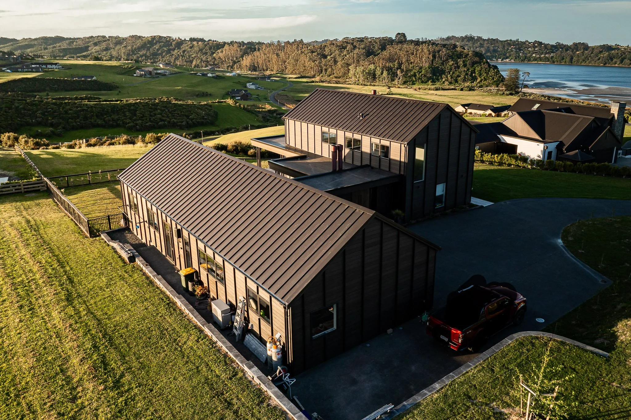 Drone perspective showing the elevated position of the home on the ridgeline, coastal forest below, and the panorama of the Hauraki Gulf.