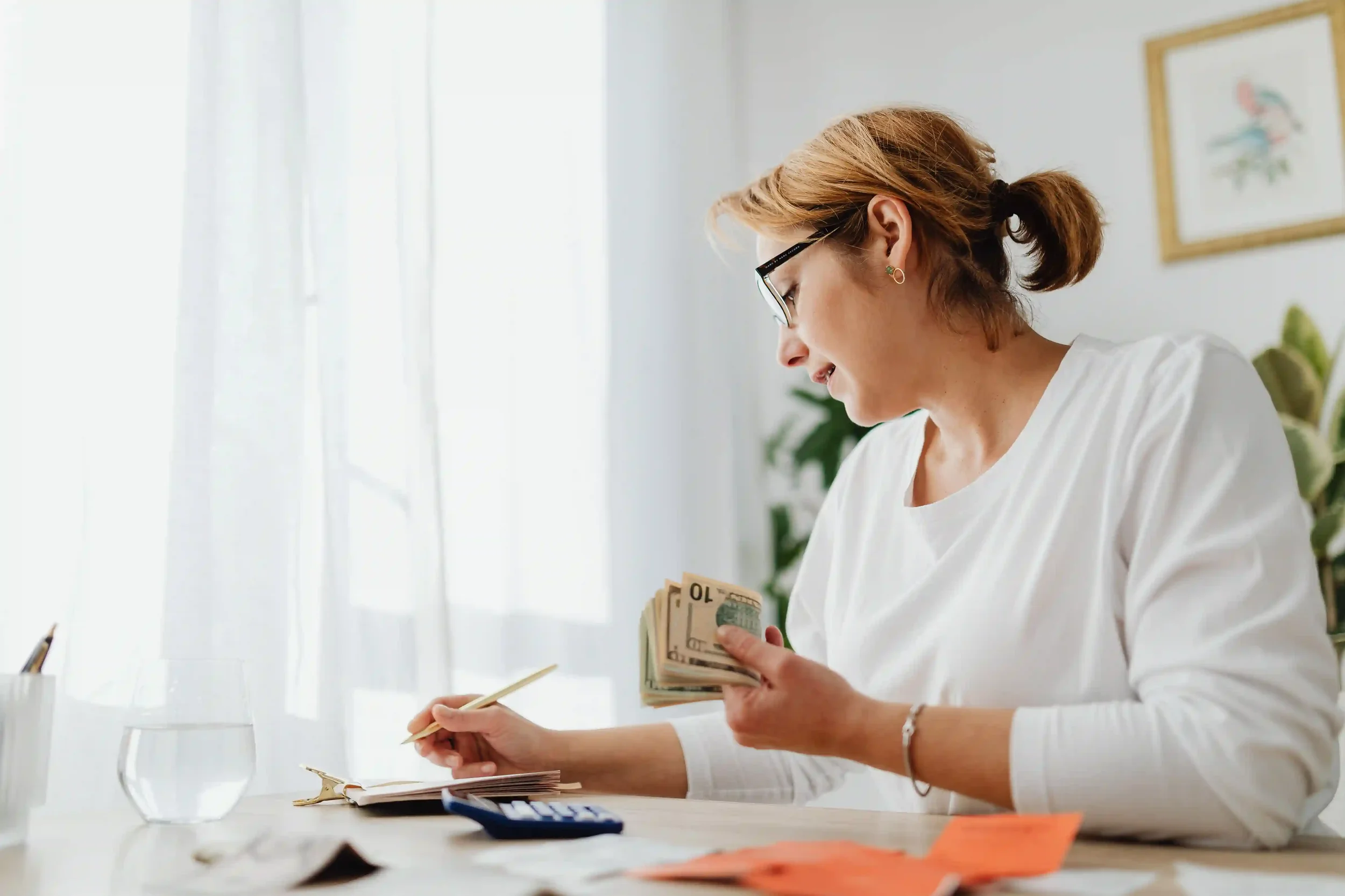 A mother reviewing expenses with a notepad and cash, budgeting for hidden costs in teen business competitions.