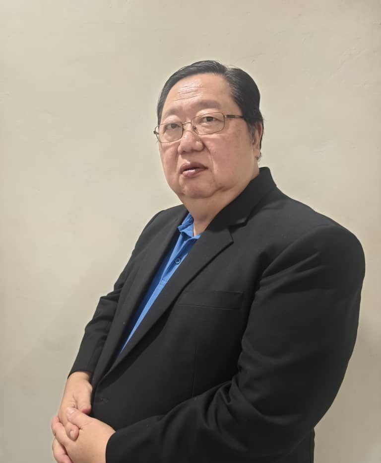 A man wearing glasses, a black blazer, and a blue polo shirt, standing against a plain beige wall, looking at the camera.