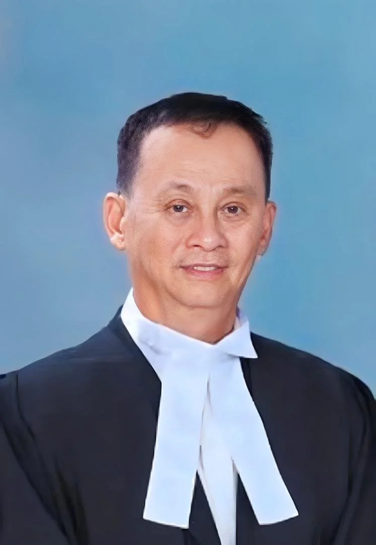 A man dressed as a lawyer or judge, wearing a black robe and white collar, against a blue background.