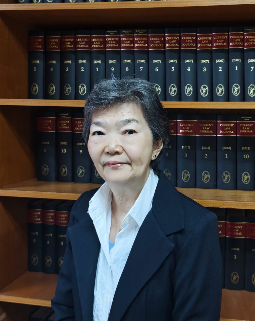 An elderly woman with short gray hair, wearing a white collared shirt and a dark blazer, standing in front of a bookshelf filled with legal books titled 'Current Law Journal'.