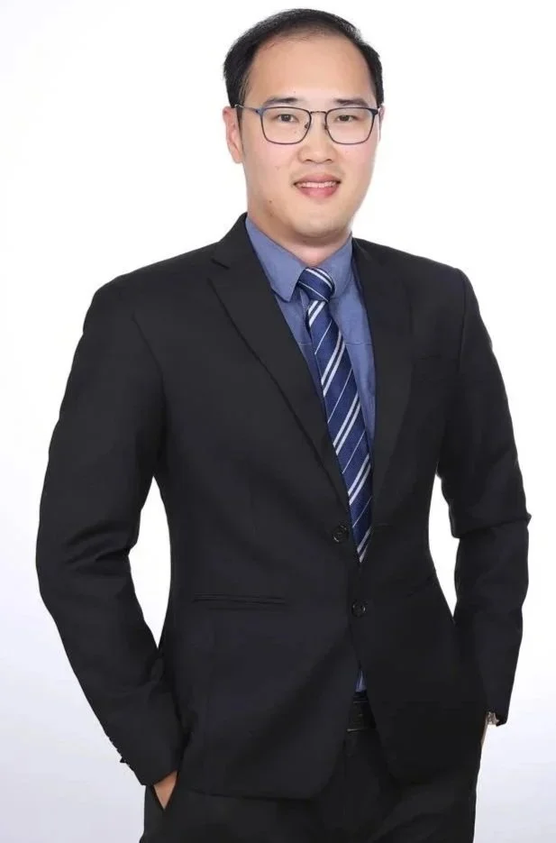 A man in a black suit, blue shirt, and striped tie standing against a white background.
