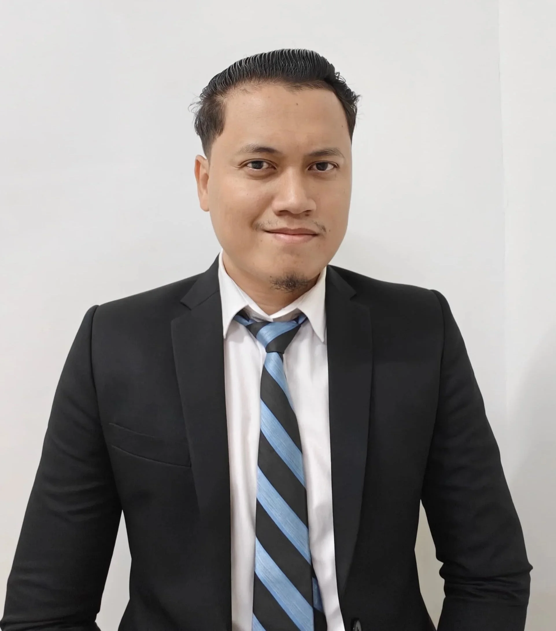 A man in a black suit, white shirt, and a blue, black, and white striped tie, smiling slightly against a plain white background.