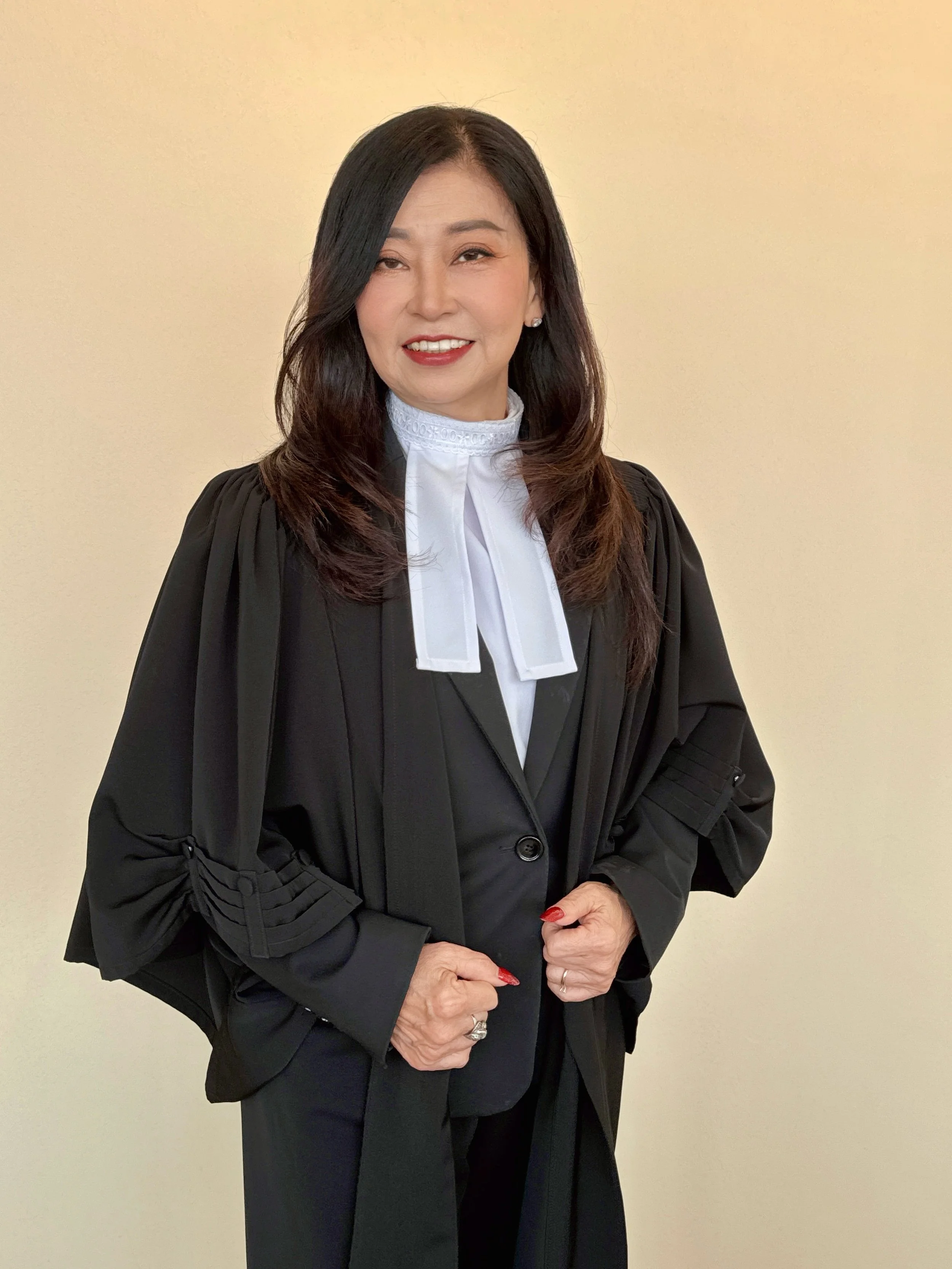 A woman dressed in a judge's robe, smiling, standing against a plain beige wall.