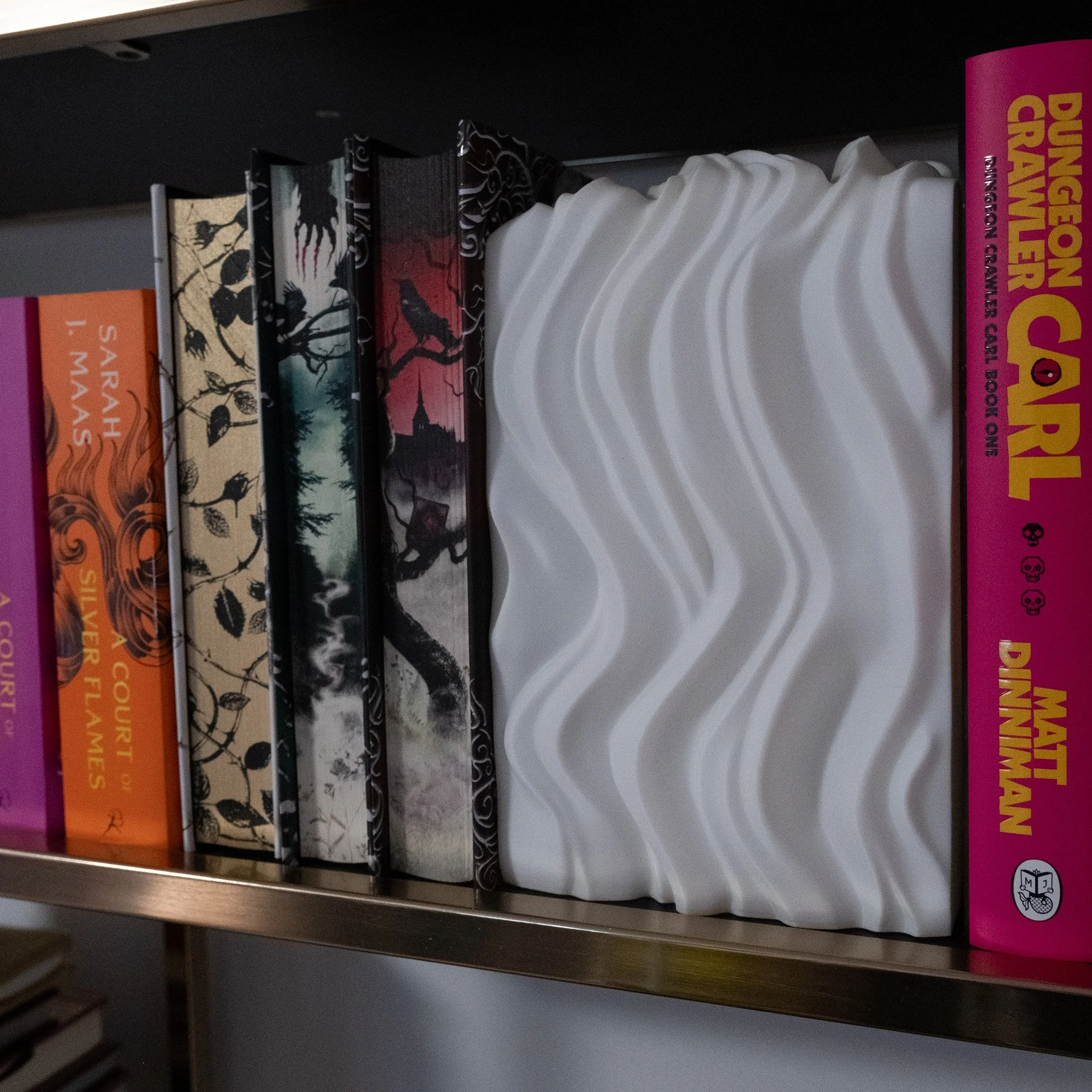 A shelf with several hardcover books and a white decorative object. The books include 'Dungeon Card Graver,' 'Matl Dinnerman,' 'Sarah I. Mas,' and several others with dark or colorful covers.