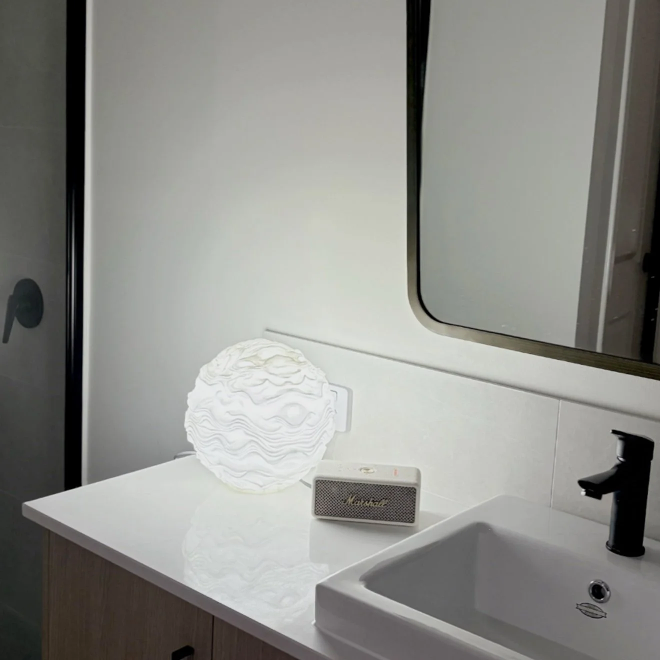 Bathroom countertop with a decorative white spherical lamp, a small Marshall speaker, and a bathroom sink with a black faucet. Mirror partially visible above the sink.