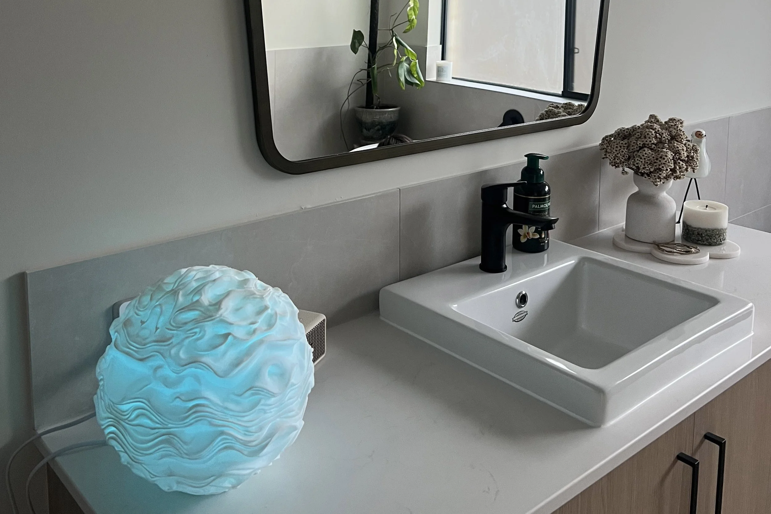 Bathroom sink with black faucet, mirror, decorative items including a vase with dried flowers, candle, and small decor pieces on a white countertop, illuminated by natural light from a window.