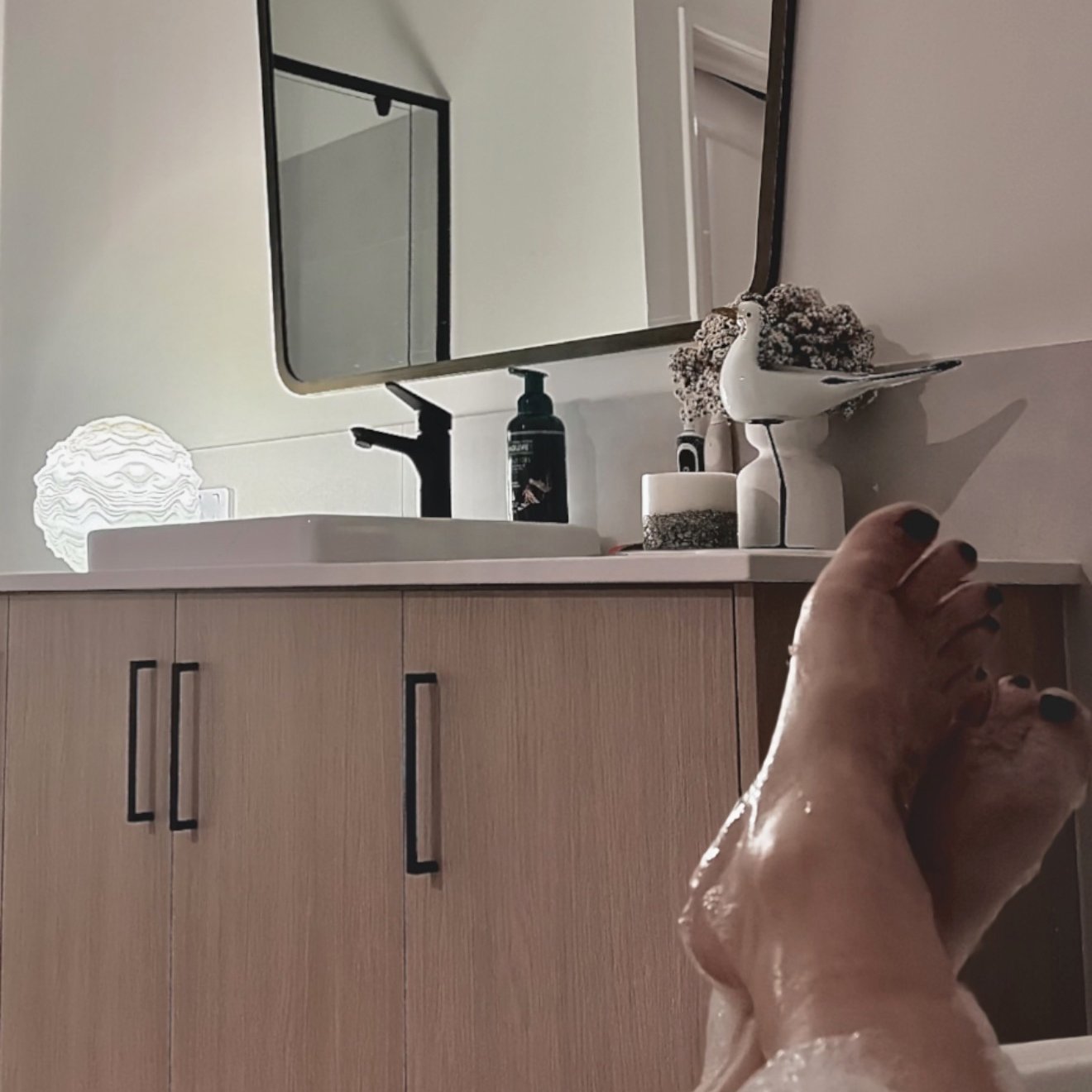 Person taking a bath, with feet visible in the foreground. The bathroom features a wooden vanity with black handles, a white sink, a black faucet, and decorative items including a white bird figurine, a jar, and some textured objects. A large mirror 