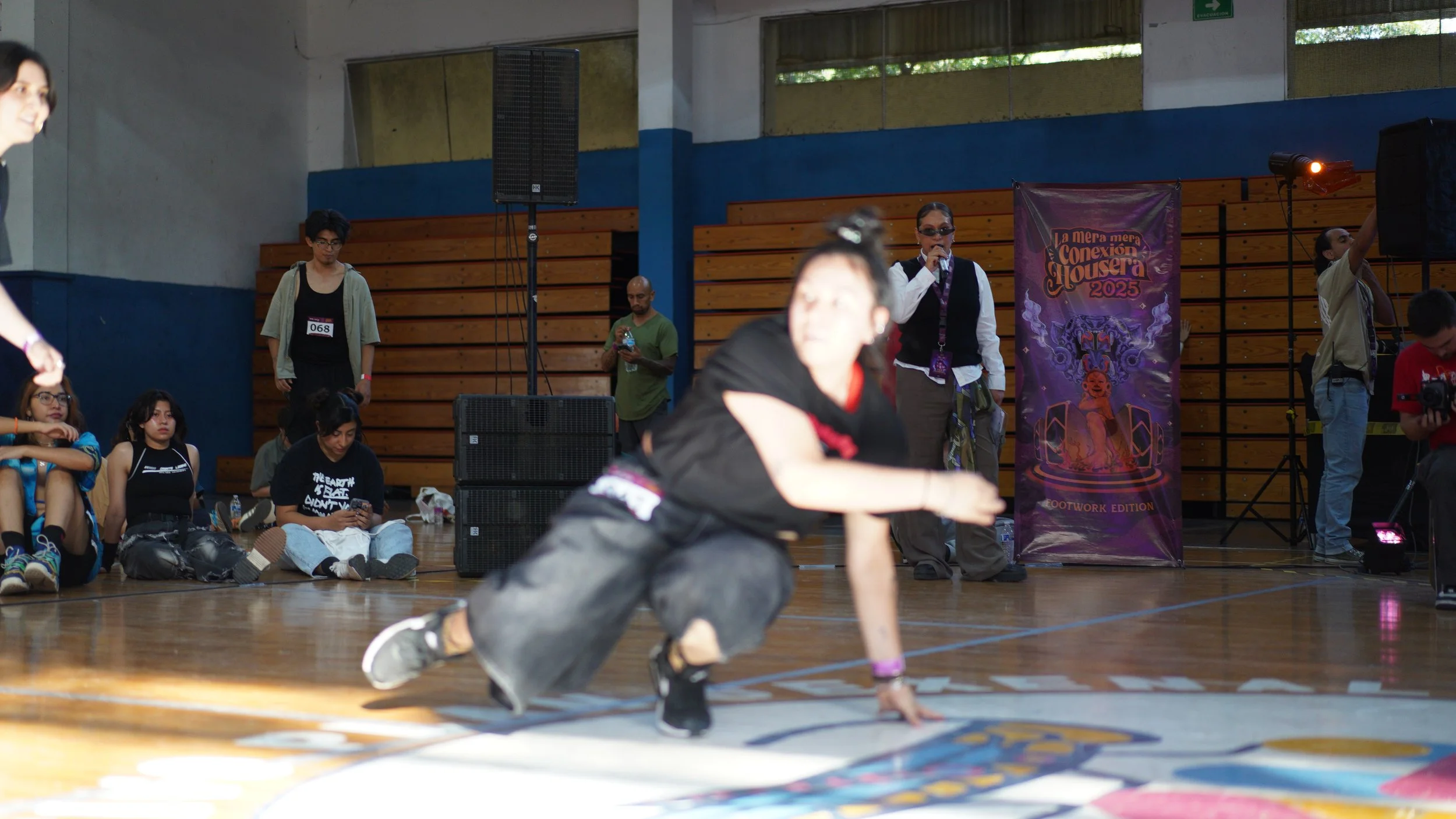 A person breakdancing on a gym floor at a dance competition with several spectators and a presenter in the background.