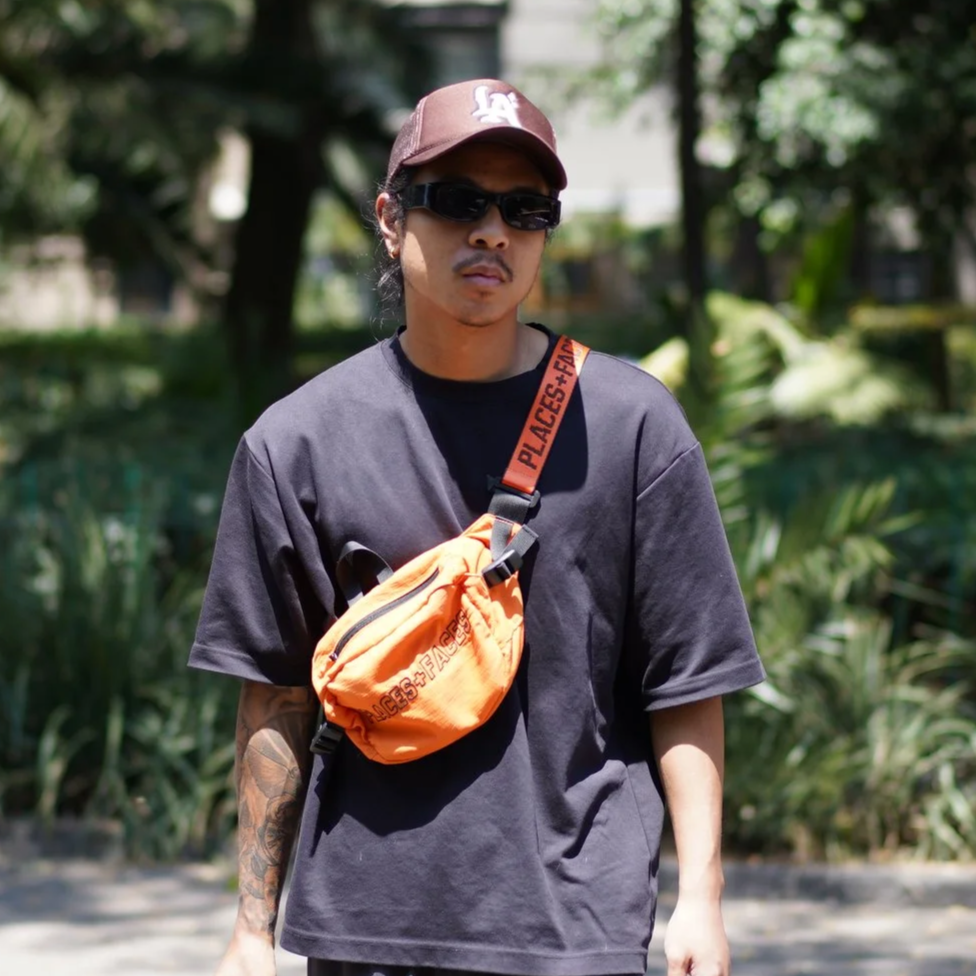 Young man in a black t-shirt, brown cap, and sunglasses walking outdoors with greenery in the background, carrying an orange crossbody bag.
