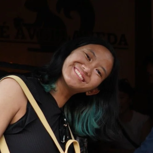 A woman with black hair and turquoise tips smiling, wearing a black top and a beige strap, indoors at a social event.