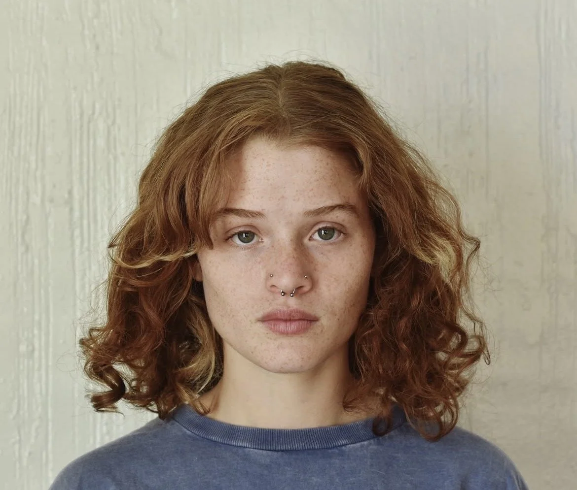 A young woman with curly red hair and a septum piercing looking at the camera against a white wall background.