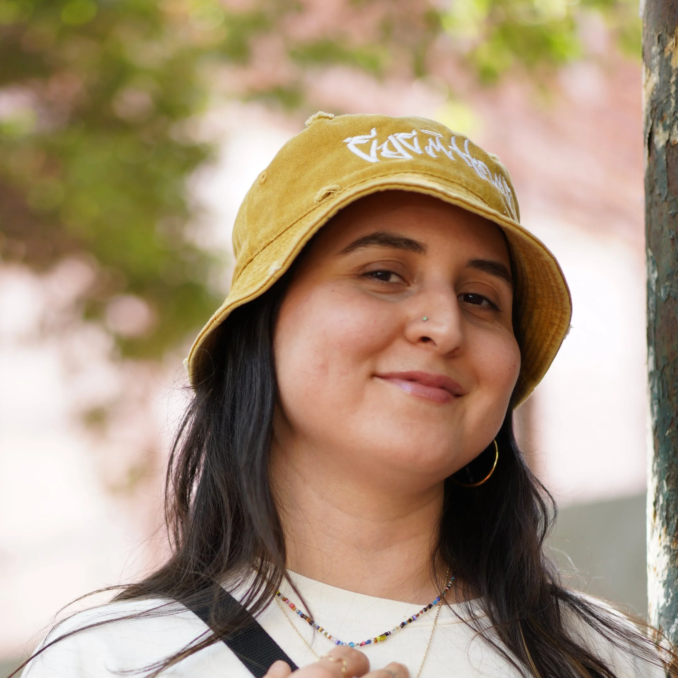Close-up of a smiling young woman with long dark hair, wearing a yellow bucket hat, small nose piercing, hoop earrings, a necklace, and a white shirt, outdoors with blurred trees in the background.
