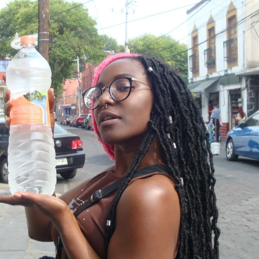 Woman with long dreadlocks, glasses, and a pink beanie holding a large bottle of water on a city street.