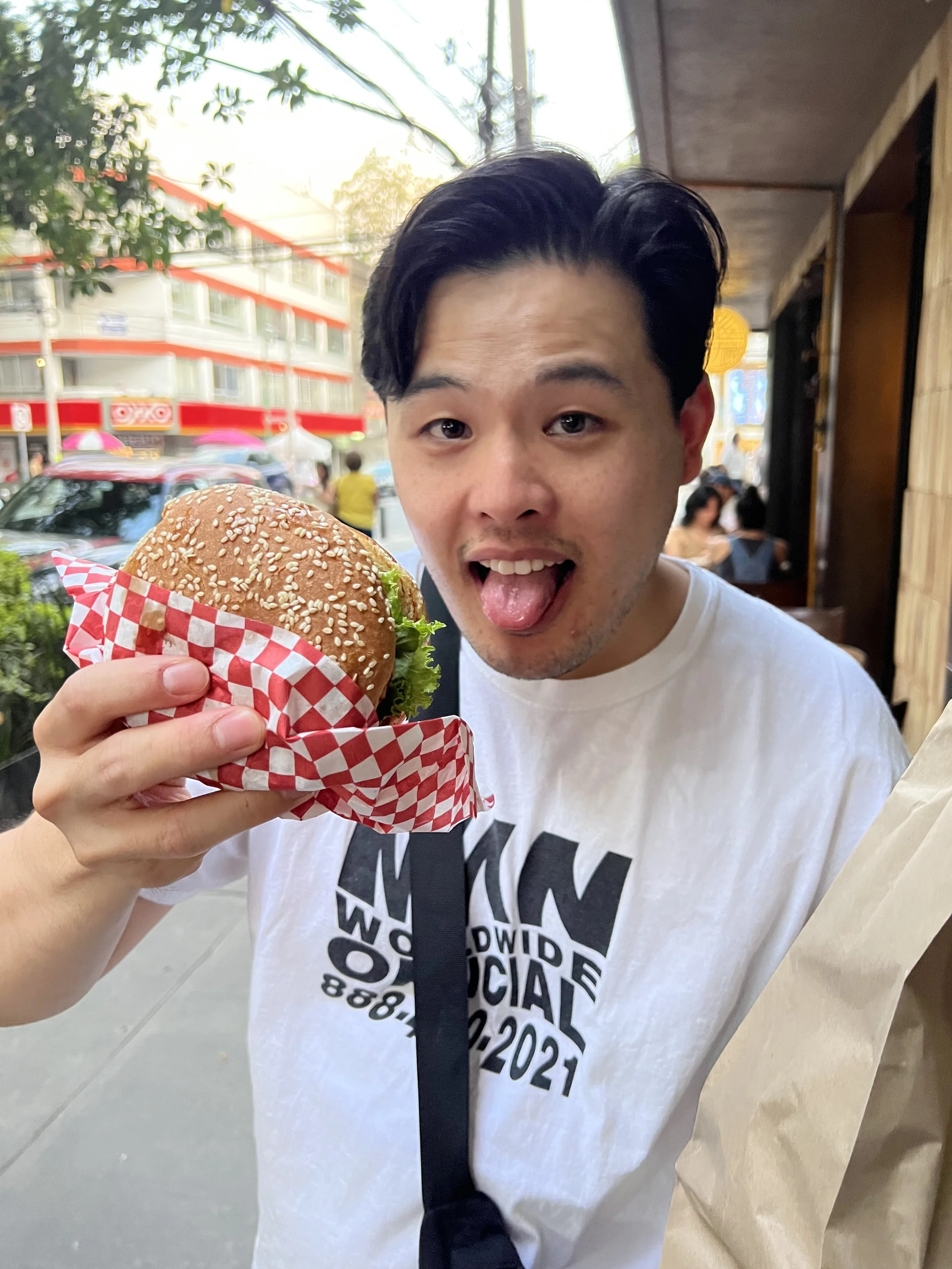 A person holding a sesame seed burger with lettuce, posing outside a restaurant with city street in the background.