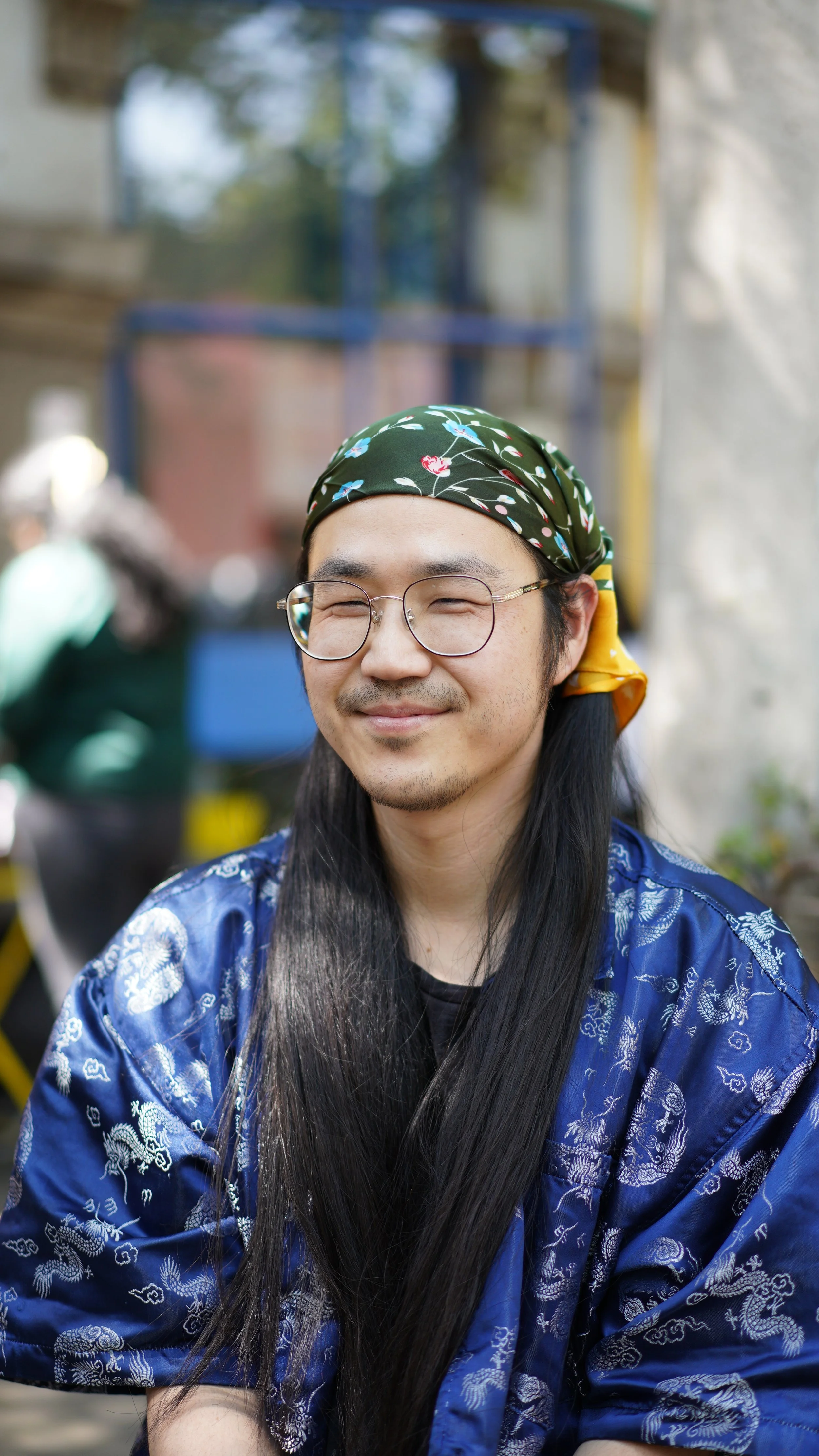 A smiling man with long black hair, glasses, wearing a green bandana with floral patterns, a black t-shirt, and a blue silk robe with white dragon designs, outdoors with a blurred background.