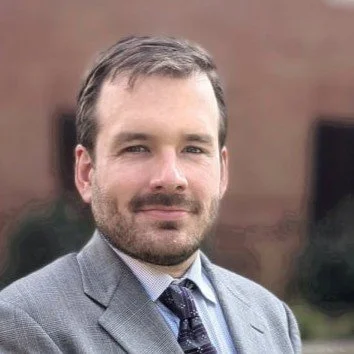A man with short brown hair and a beard wearing a gray suit and a plaid tie, standing outdoors with a blurry brick building in the background.