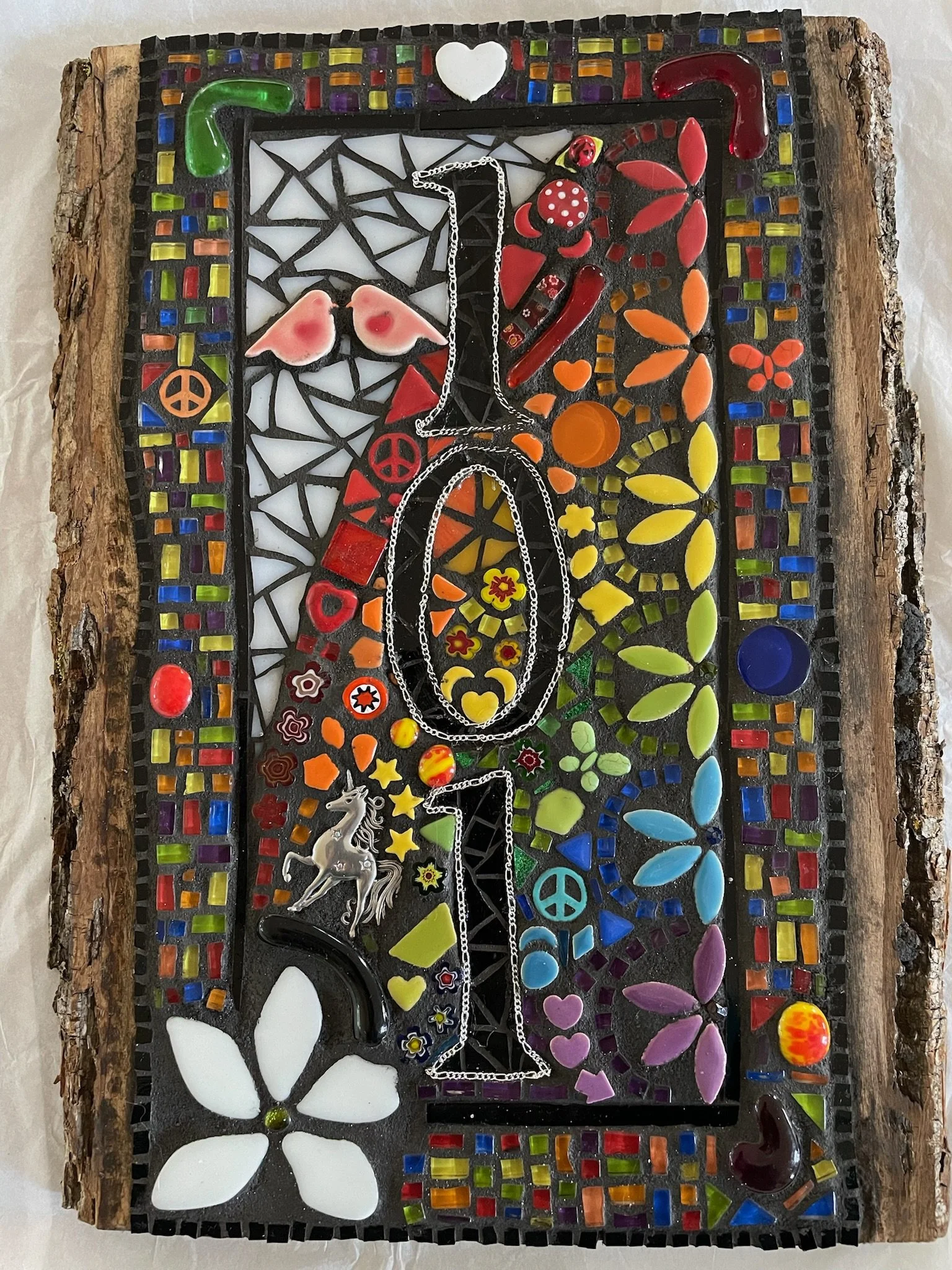 Colorful mosaic artwork featuring peace symbols, flowers, hearts, rainbows, a unicorn, and jewelry with a black wood border.