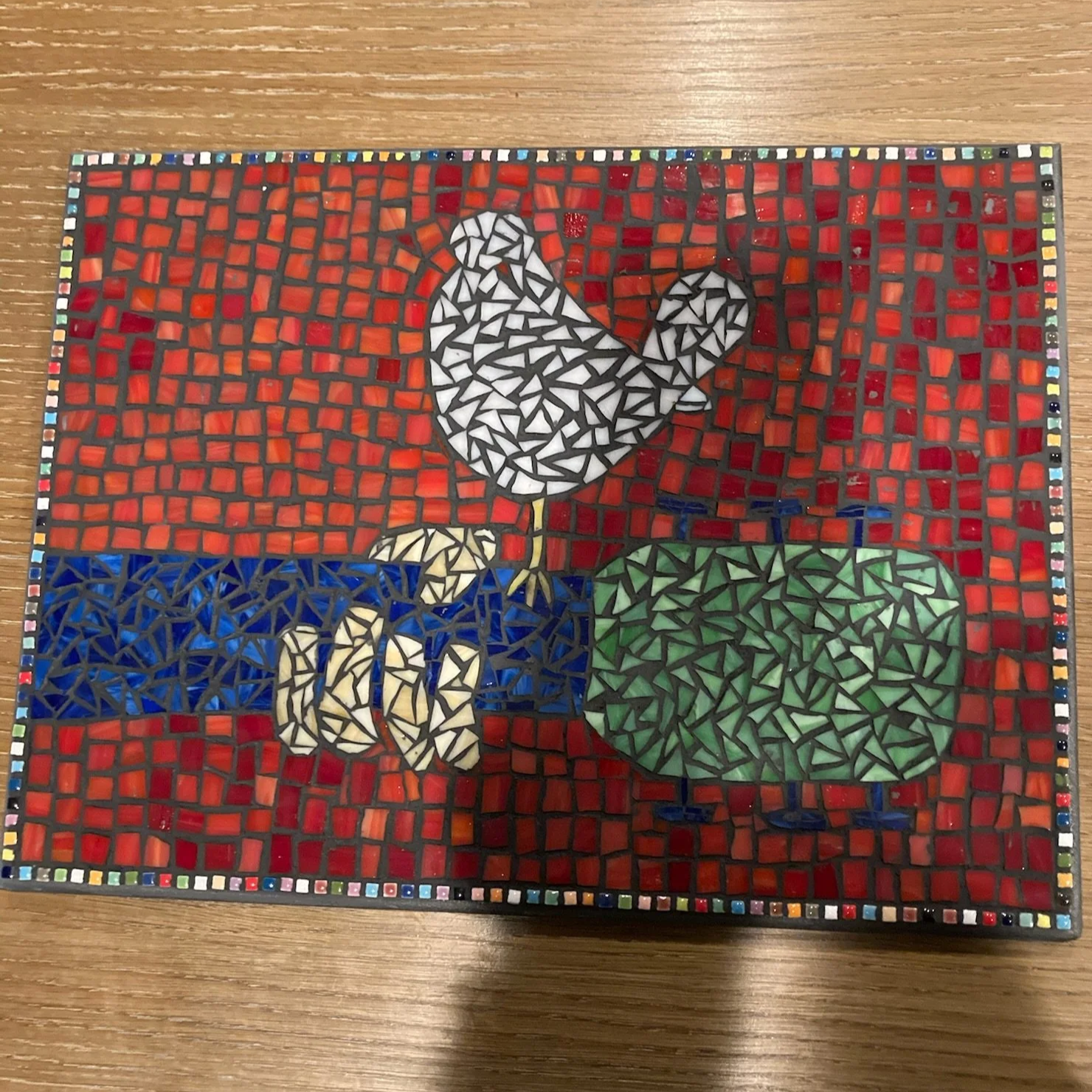 Decorative mosaic artwork of a chicken with white and black feathers, standing on one leg beside a green vegetable, set against a red and blue background.