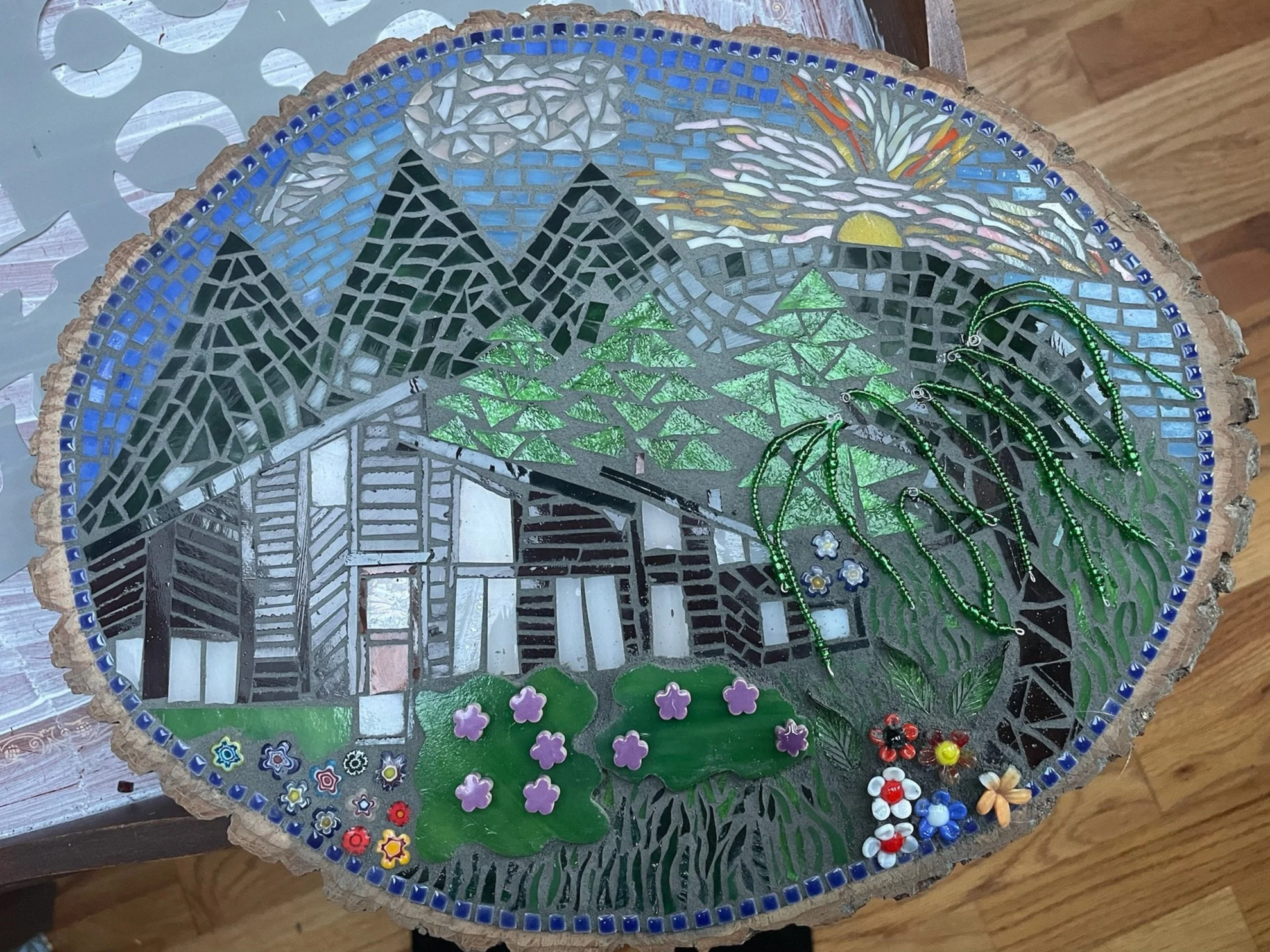 Mosaic art of a mountain landscape with a cabin, green trees, a bright sun, a rainbow, and flowers, all set on a wood slice table.