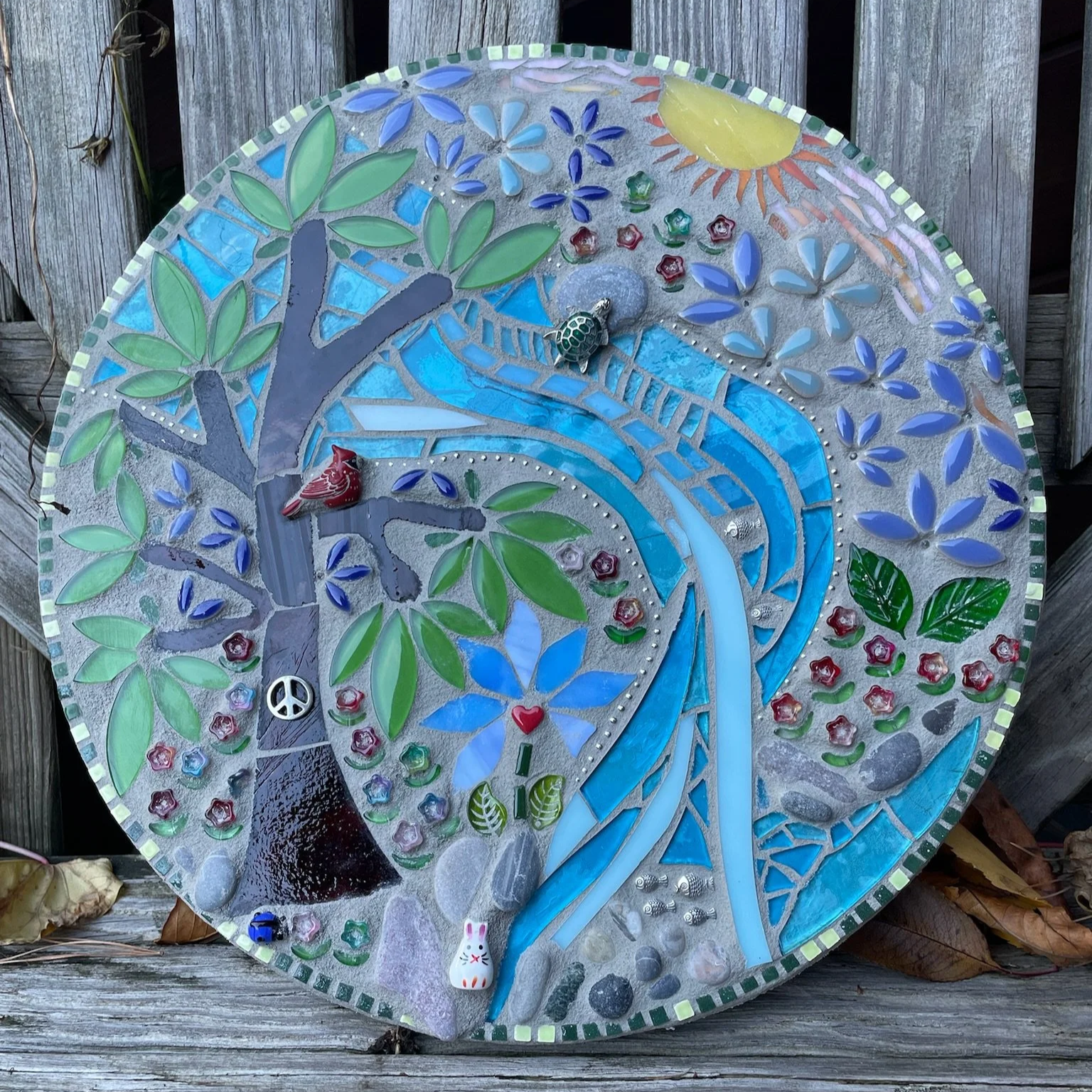 Colorful mosaic clock featuring a flowing blue river, green trees, flowers, a bird, a sun, and decorative objects, mounted outdoors.