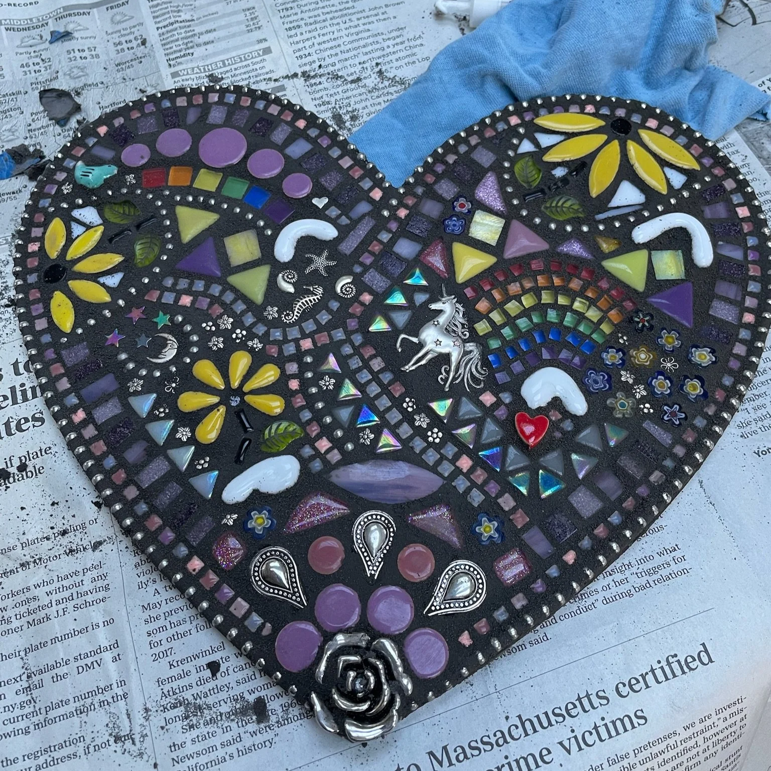 Colorful mosaic heart with various decorative elements including a unicorn, rainbow, flowers, crescent moons, and hearts, placed on newspaper.