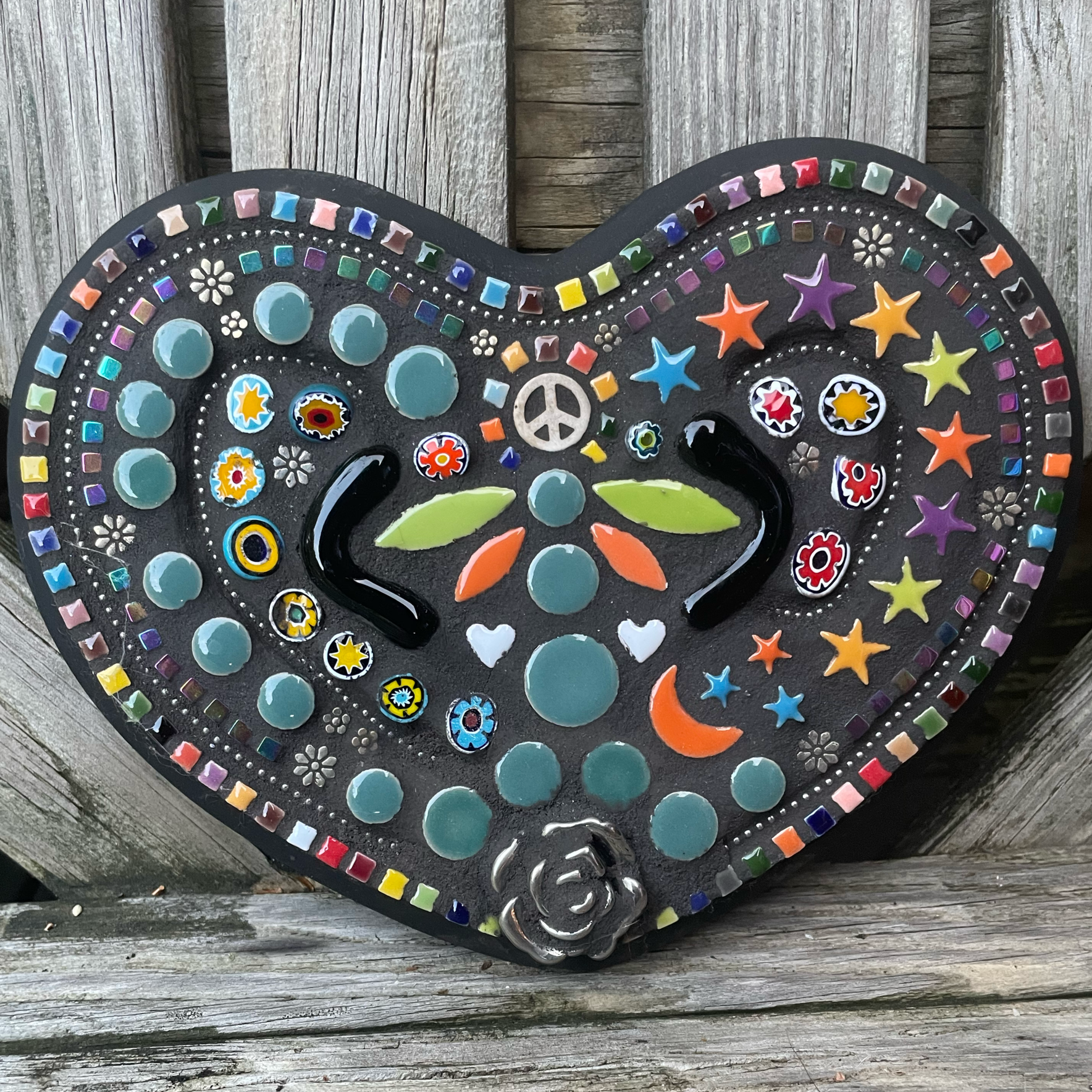 Colorful mosaic heart-shaped wall art featuring stars, moons, peace sign, and floral patterns, mounted on weathered wood.