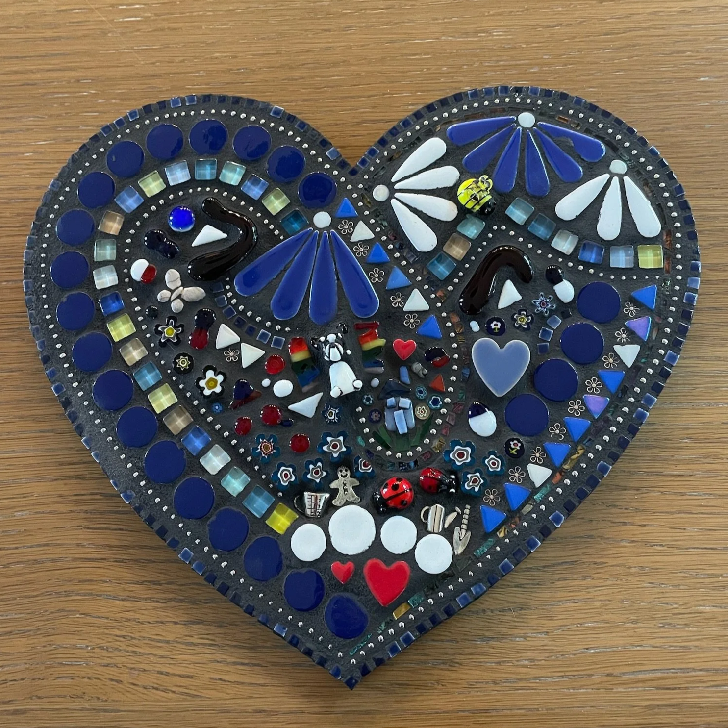 Heart-shaped mosaic artwork made of various glass and ceramic pieces, including flowers, hearts, and abstract shapes in blue, white, black, red, and other colors, on a wooden surface.