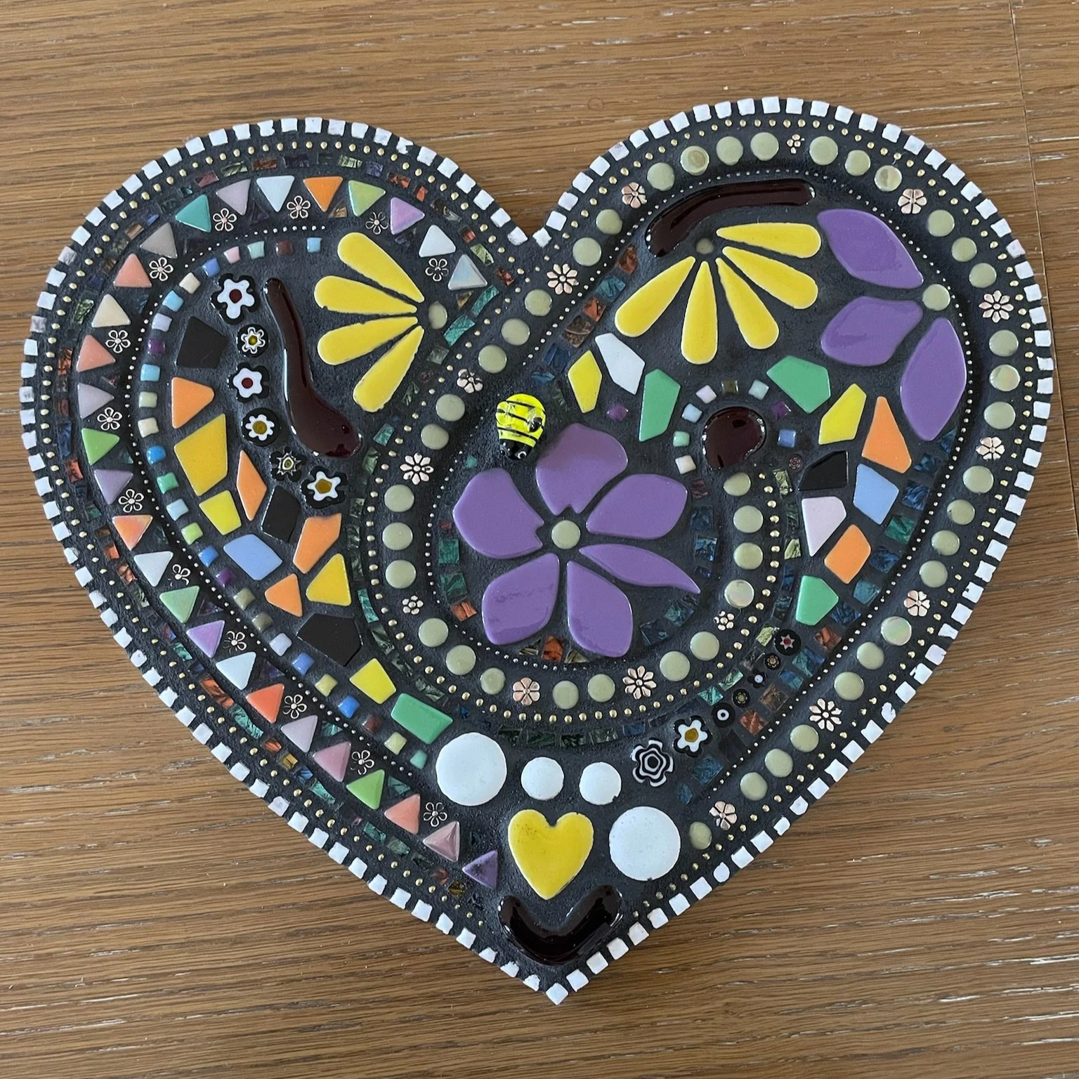 Heart-shaped mosaic tray decorated with colorful ceramic tiles, beads, and floral patterns, featuring purple and yellow flowers, a small bee, and various geometric designs on a wooden surface.