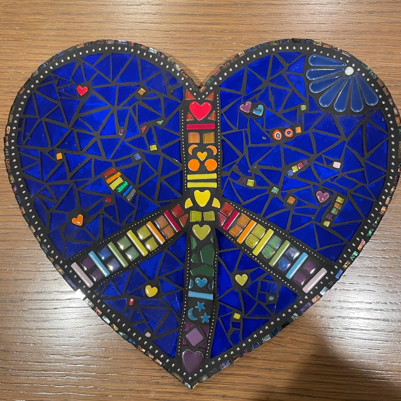 Mosaic heart-shaped art piece with rainbow colors and various small decorations on a wooden surface.