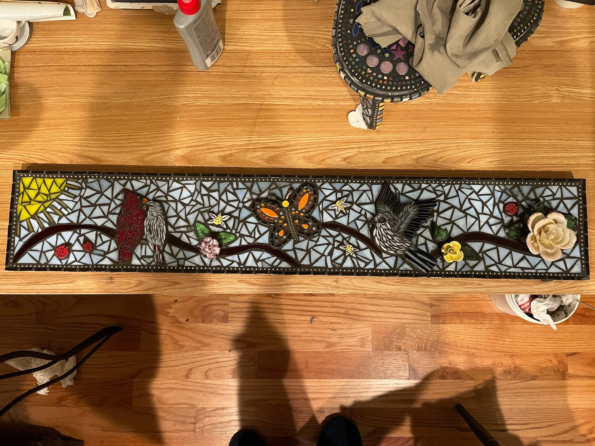 A long rectangular mosaic art piece featuring birds, butterflies, flowers, ladybugs, and a sun, all set against a background of light blue mosaic tiles with black borders.