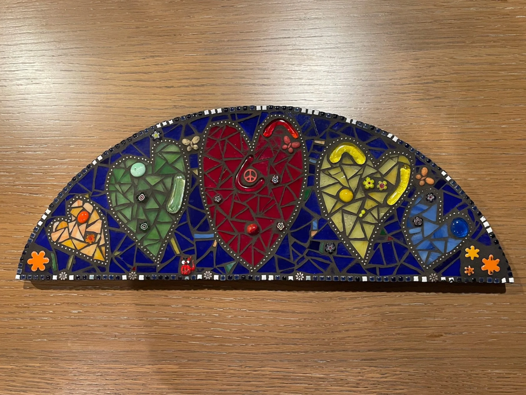 Colorful mosaic art piece with five hearts in different colors and various small decorative elements on a wooden surface.