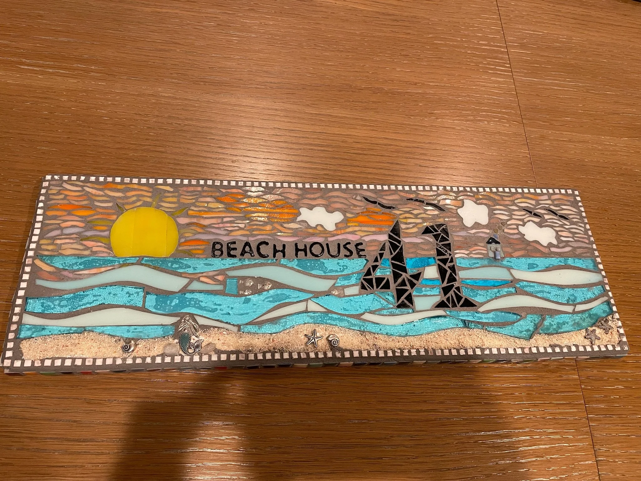 Decorative mosaic sign with a beach scene, including a yellow sun, blue waves, a rock formation, and a lighthouse. The sign reads "Beach House."