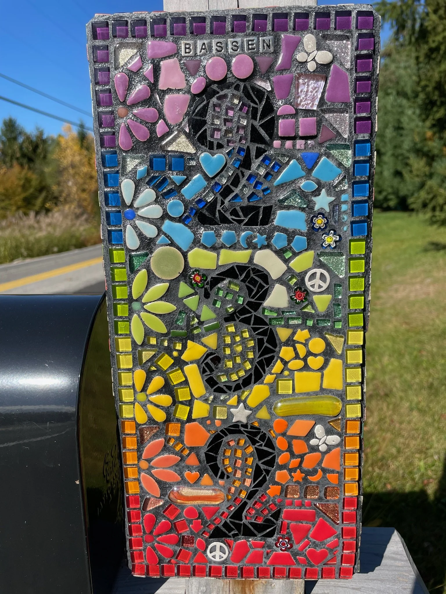Colorful mosaic art piece featuring the word 'BASSEN' at the top, with a black silhouette of a person holding a cup or glass in the center, surrounded by various geometric shapes, flowers, hearts, peace signs, and stars arranged vertically in section
