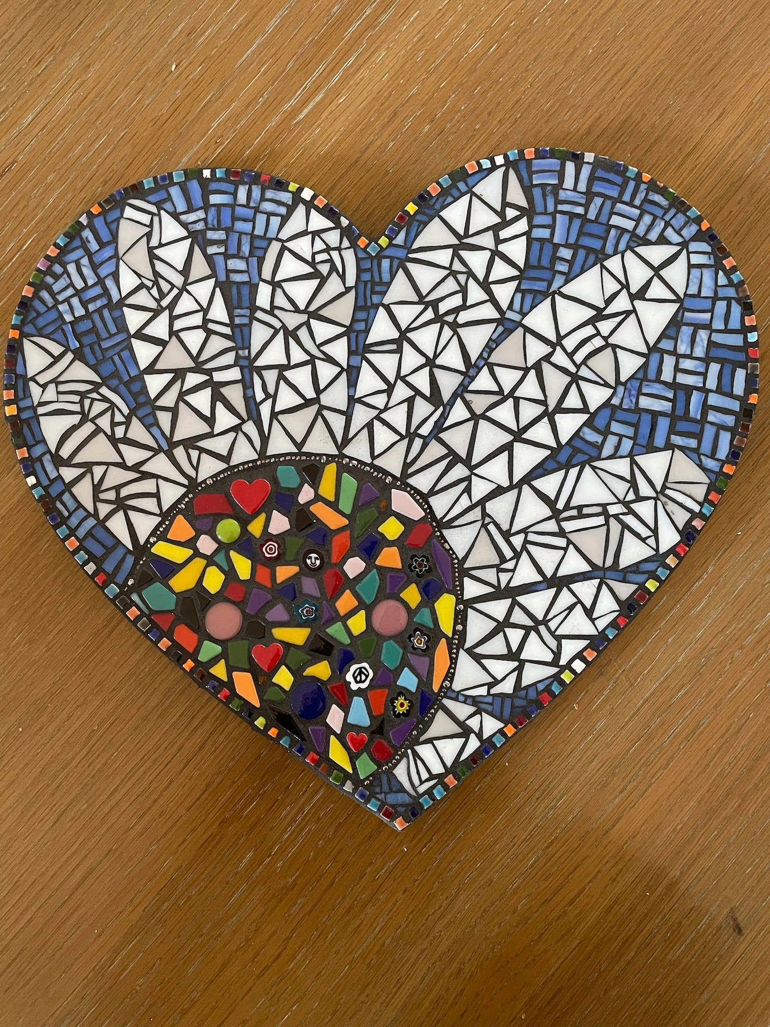 Mosaic heart-shaped piece with a multicolored skull design in the center, surrounded by white, blue, black, and multicolored tiles.