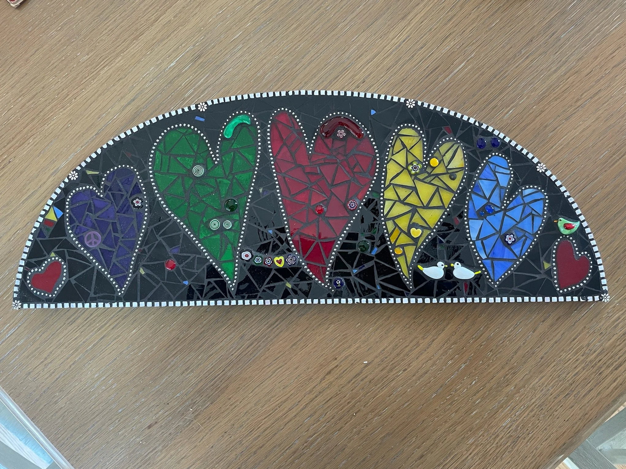 Colorful mosaic with six large hearts in purple, green, red, yellow, blue, and red, all outlined with dots. The background is black with small dots and decorative elements including small hearts, peace signs, and musical notes. Two white birds are ne