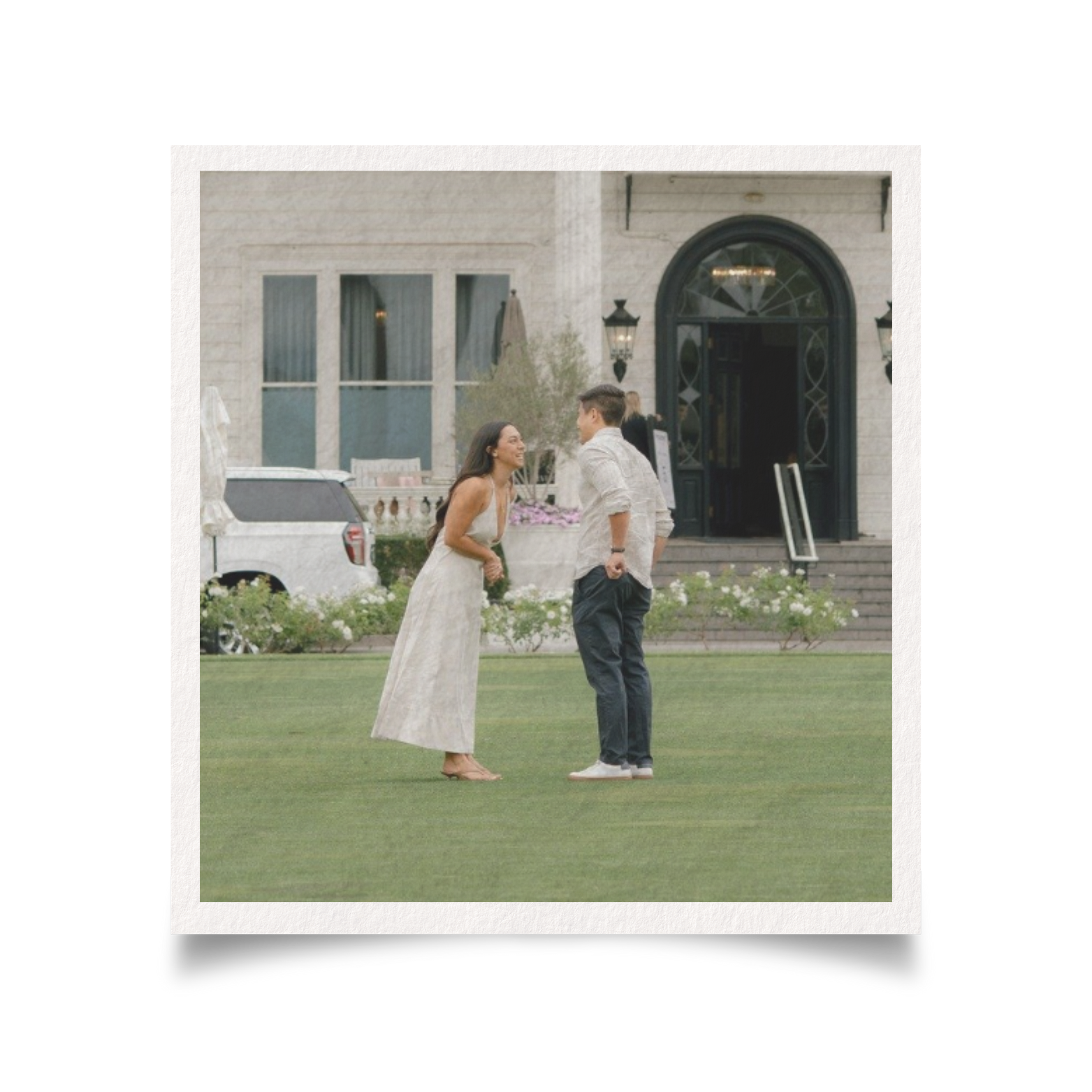 A woman in a white dress and a man in a beige shirt with rolled-up sleeves and dark pants are outside on a lawn, smiling and looking at each other, with a large building in the background.