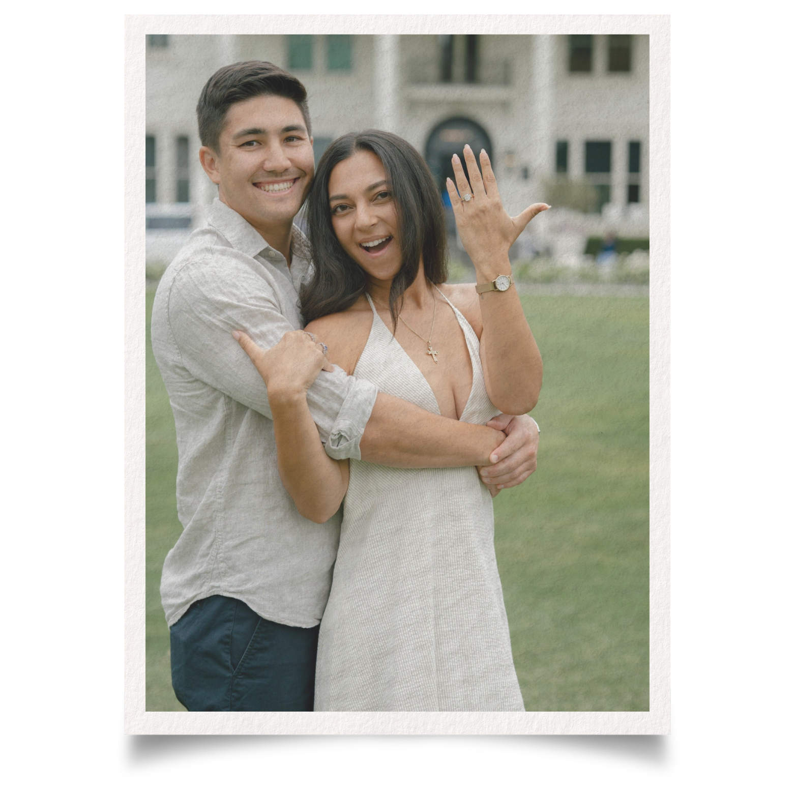 A happy couple outdoors, the man hugging the woman from behind, the woman showing her engagement ring and smiling, set against a background of a house and yard.